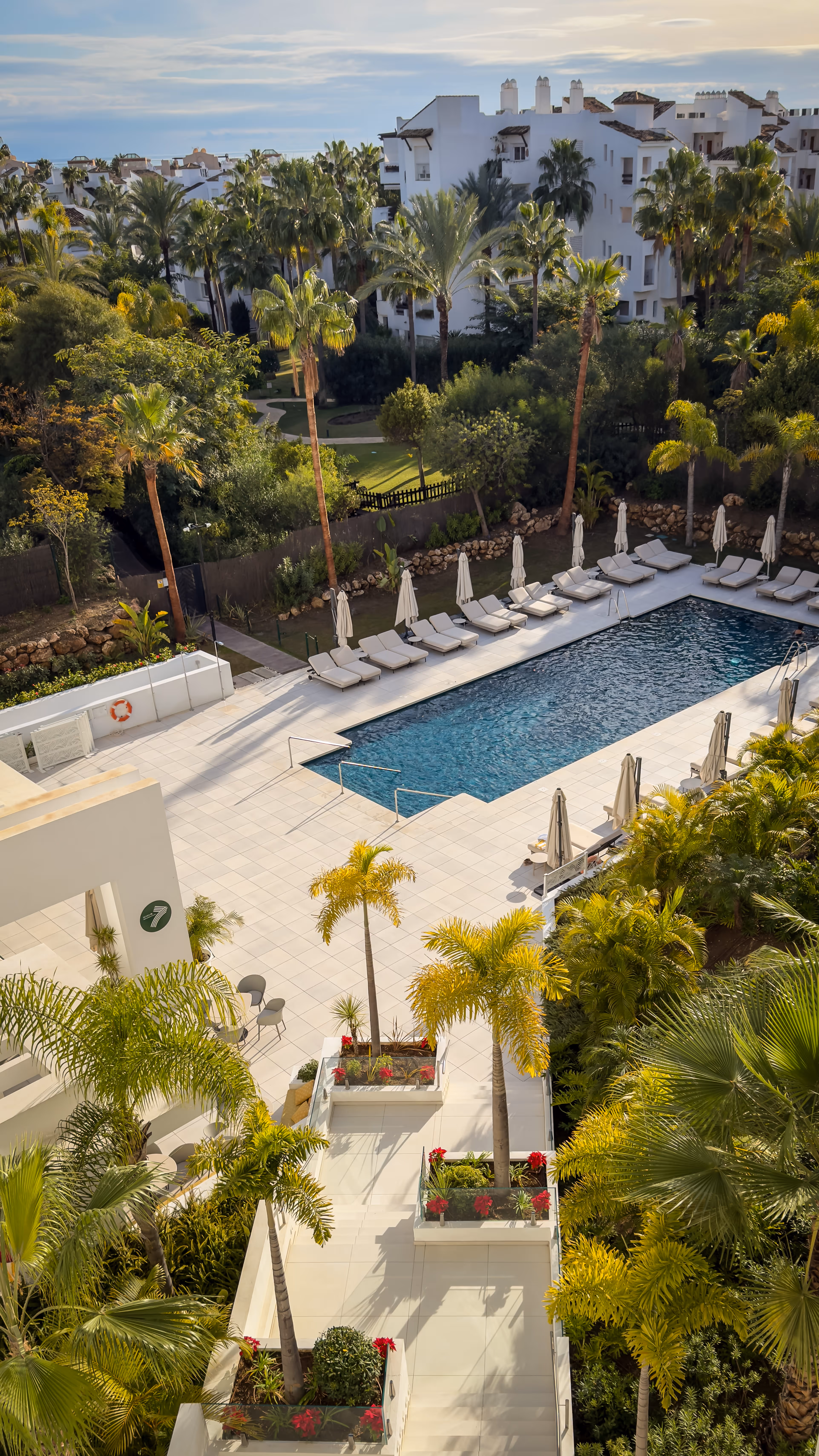 Outdoor pool area with white lounge chairs and umbrellas surrounded by palm trees and white residential buildings.
