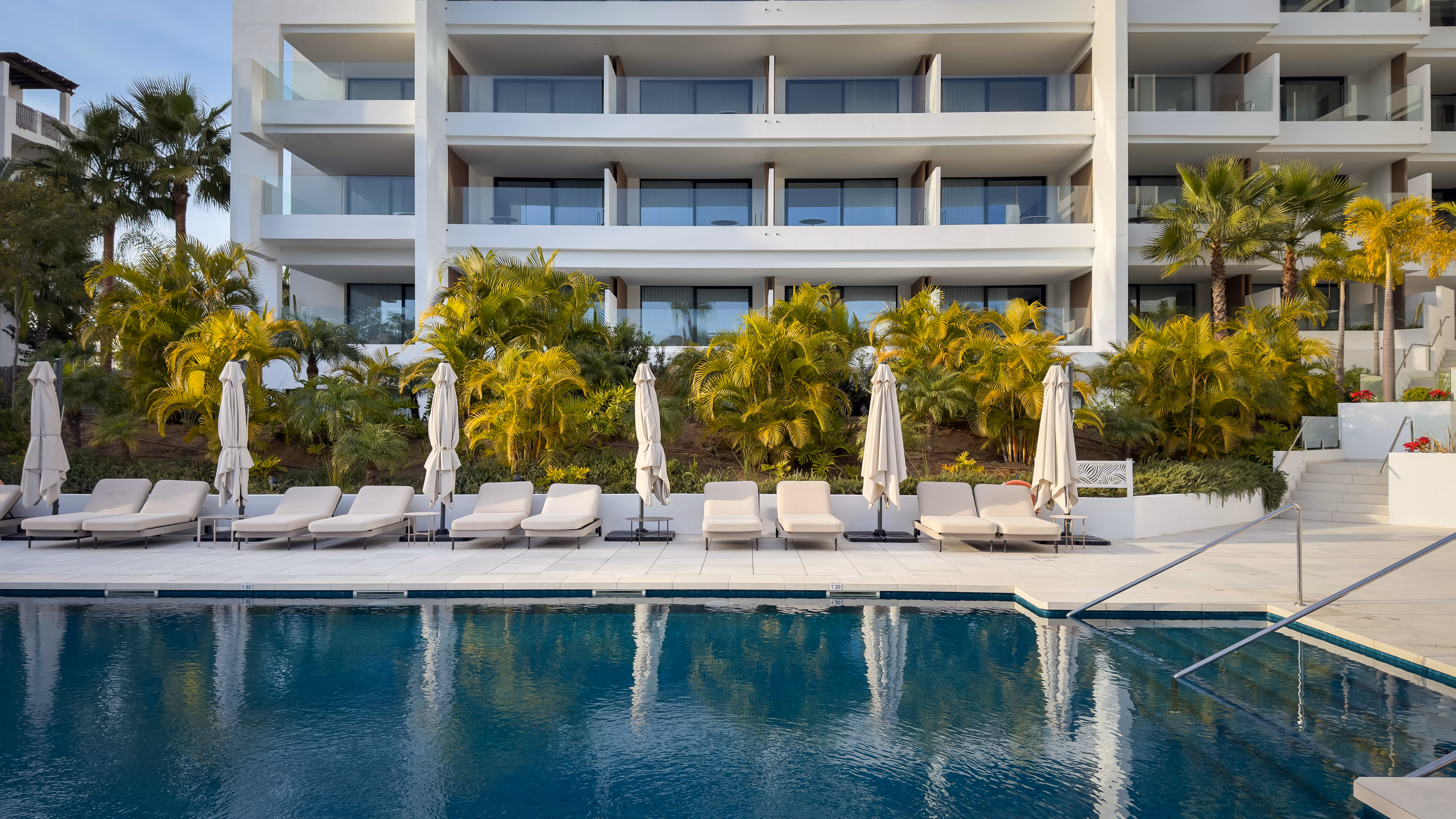 Outdoor pool beside green lawn with red flowering bushes, lounge chairs, and mountainous forest background under clear blue sky.