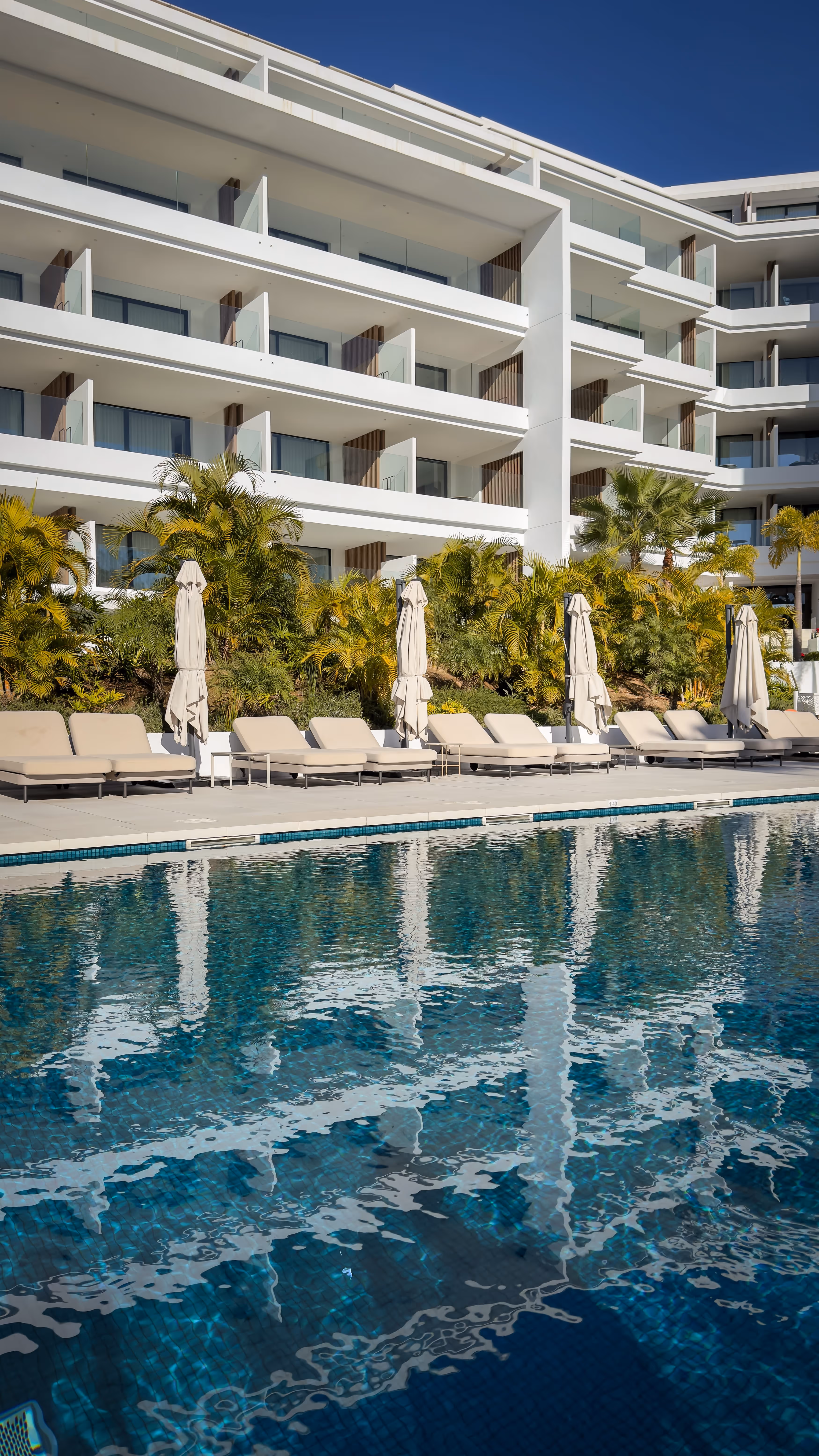 Outdoor swimming pool with reflections of a modern white apartment building and beige lounge chairs under closed umbrellas.