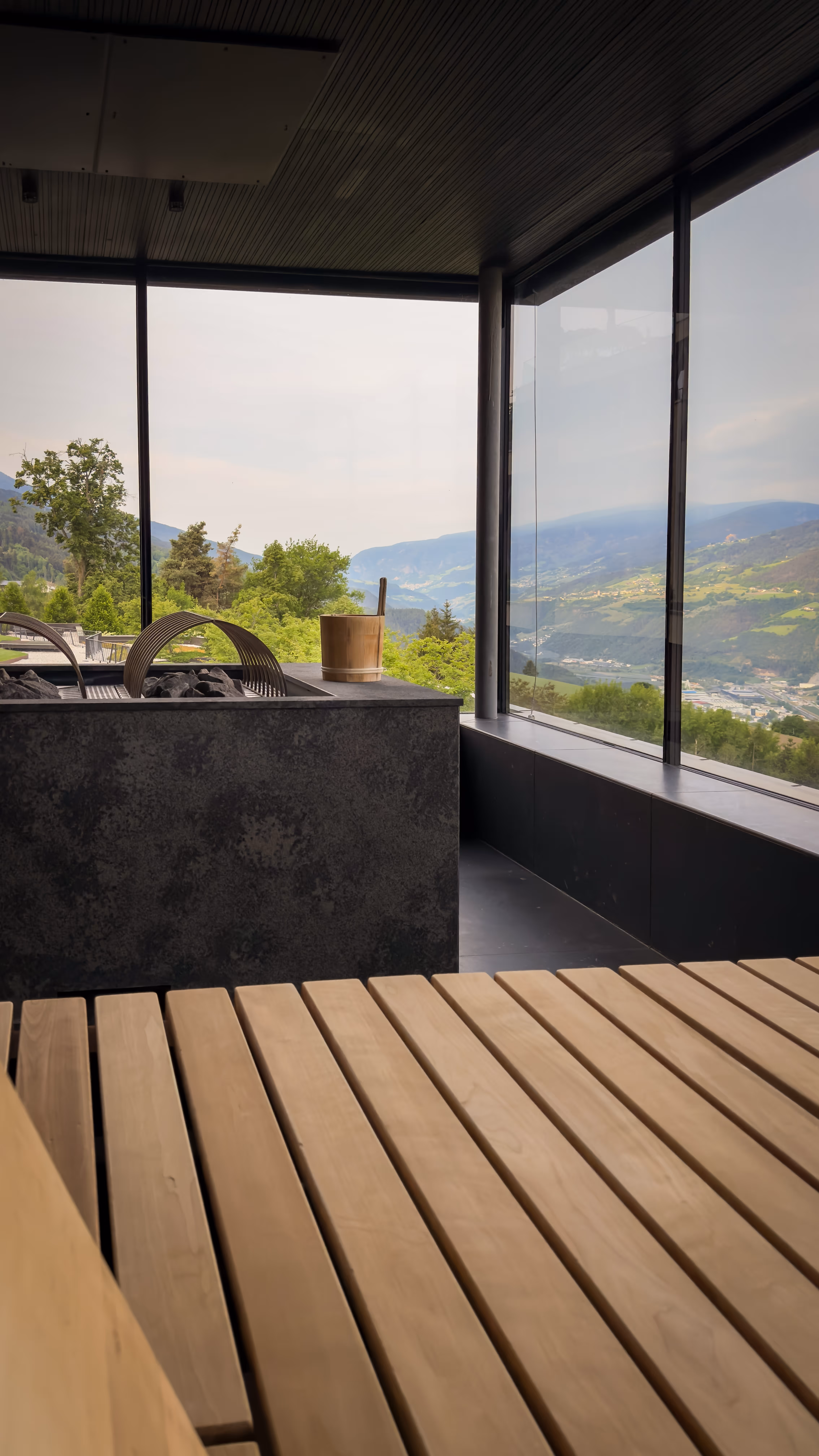 Modern sauna interior with wooden benches, black stove, and large windows showing mountain and valley views.