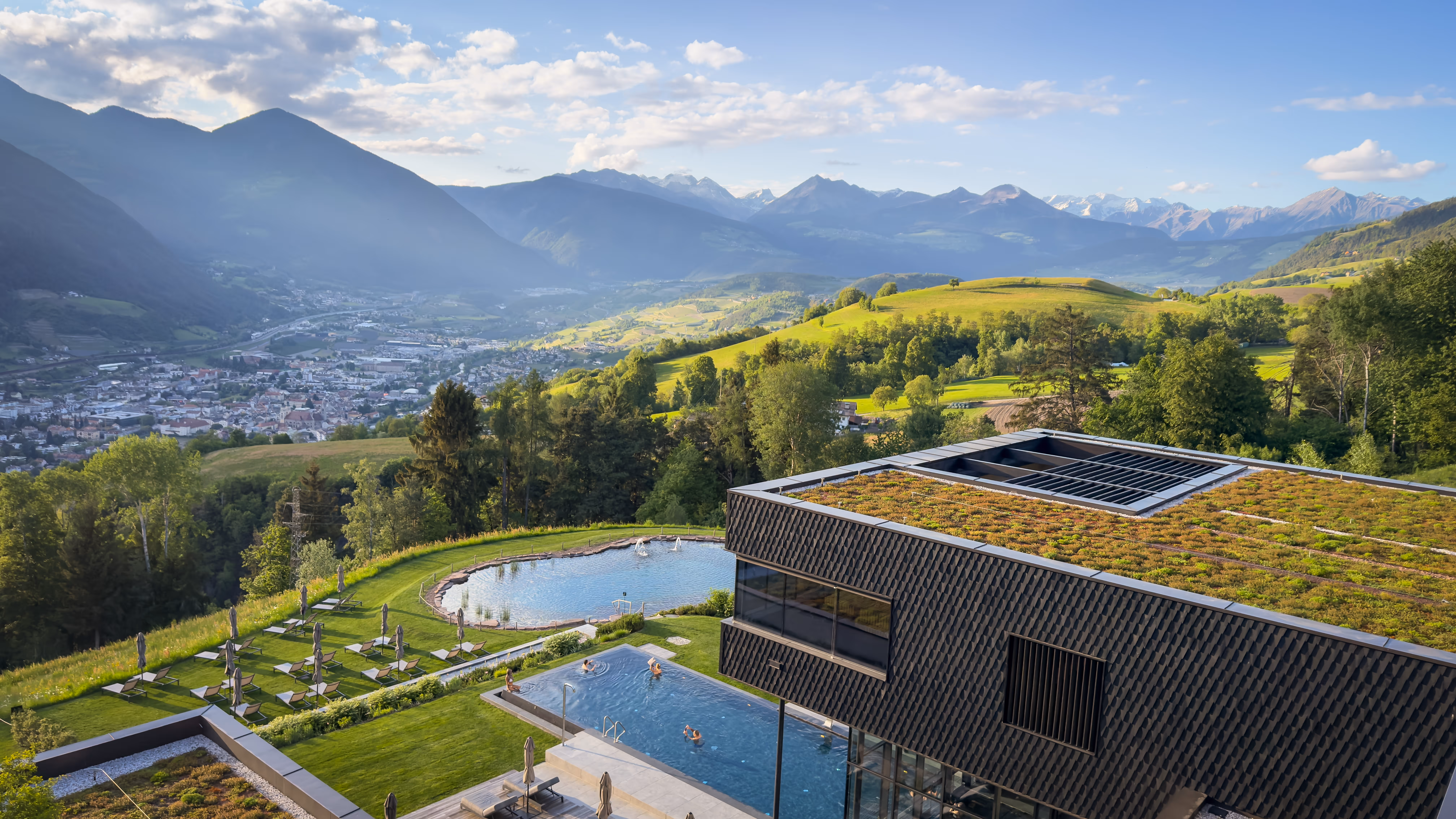 Outdoor pool with sun loungers, red flowers, green lawn, trees, and forested mountains in background.