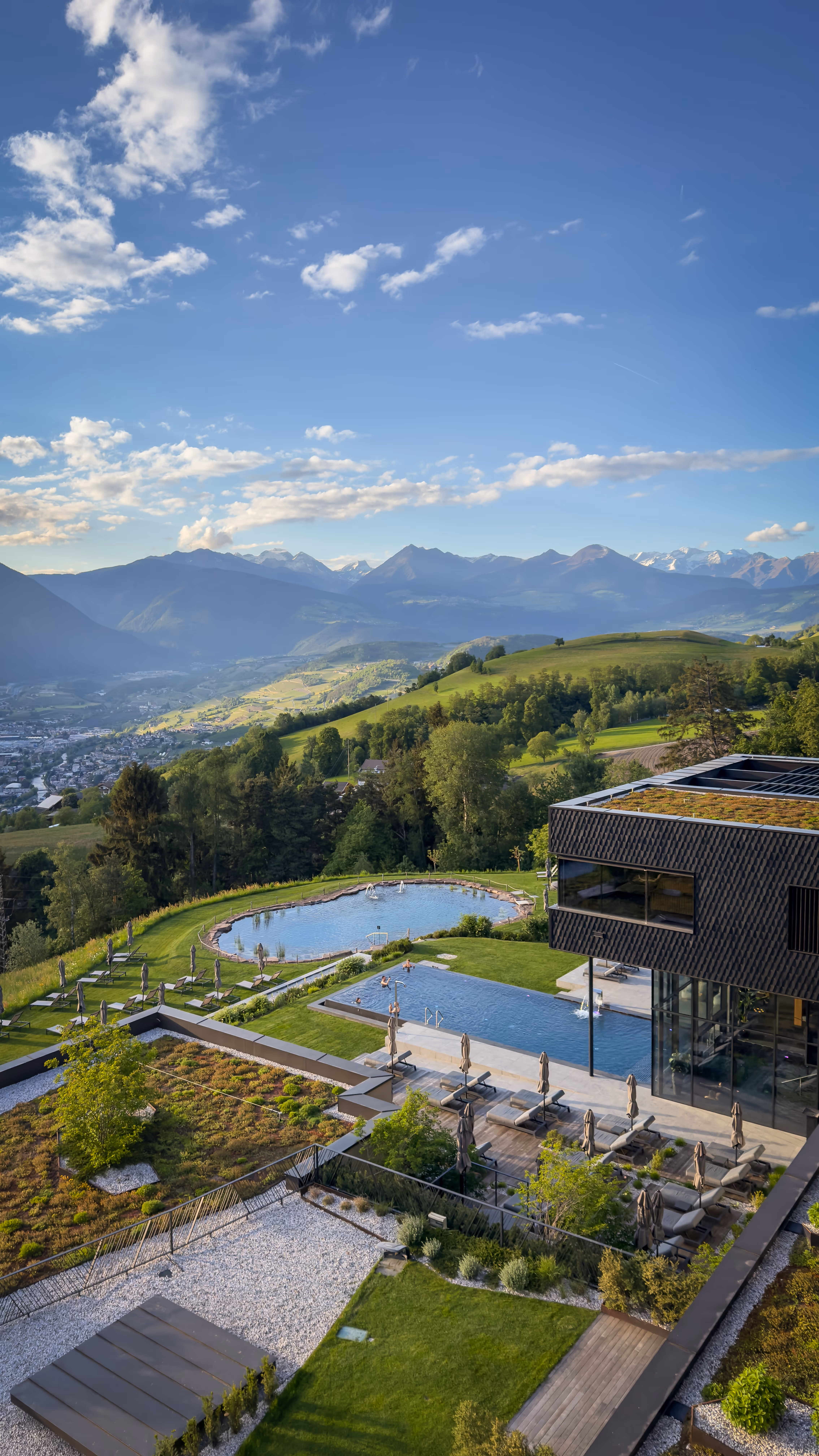 Outdoor pool with a small arched wooden bridge, surrounded by lounge chairs, greenery, and a hotel building with mountains in the background under clear blue sky.