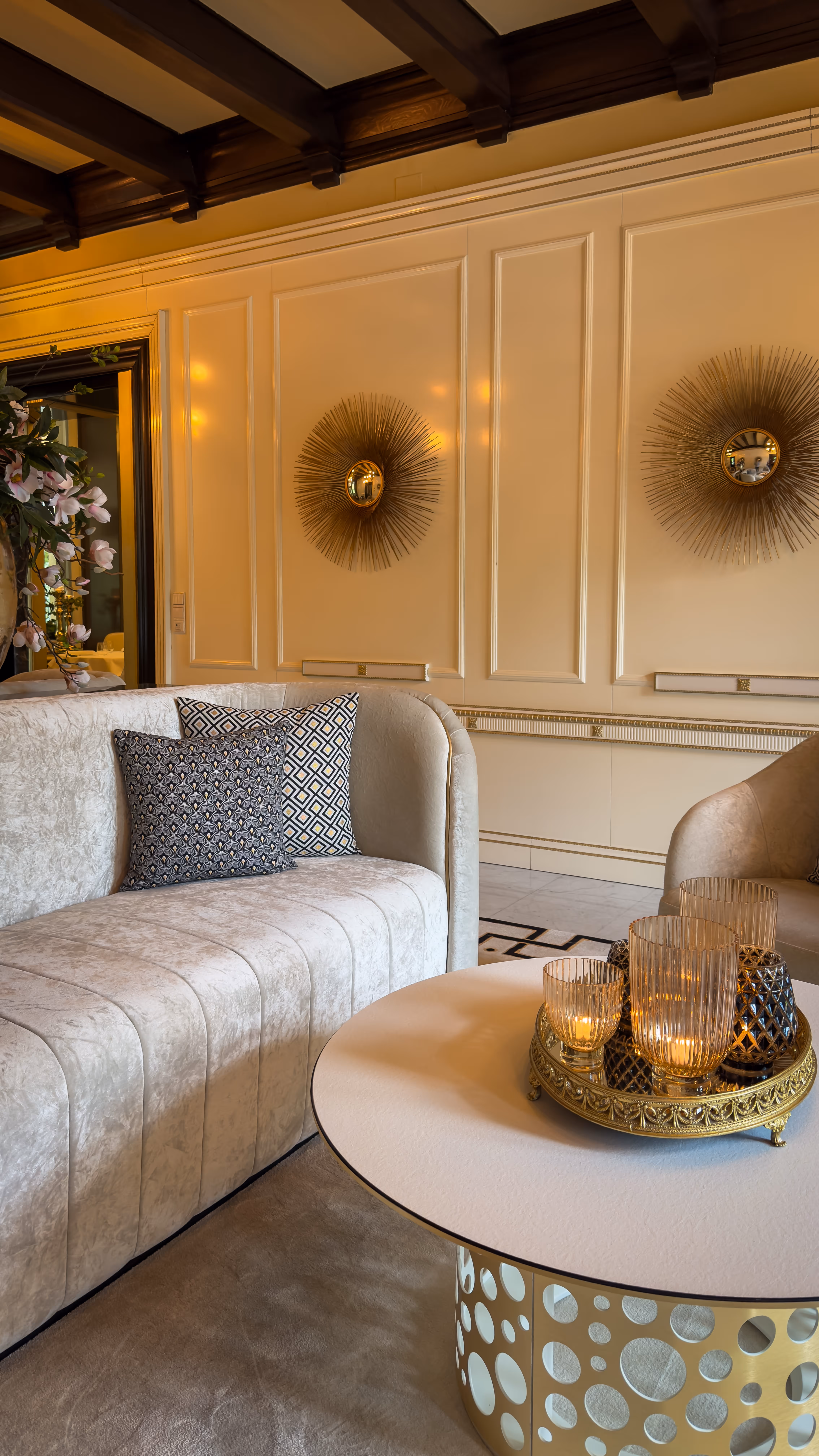 Elegant living room with beige velvet sofa, patterned pillows, round table with candles, and decorative wall mirrors.