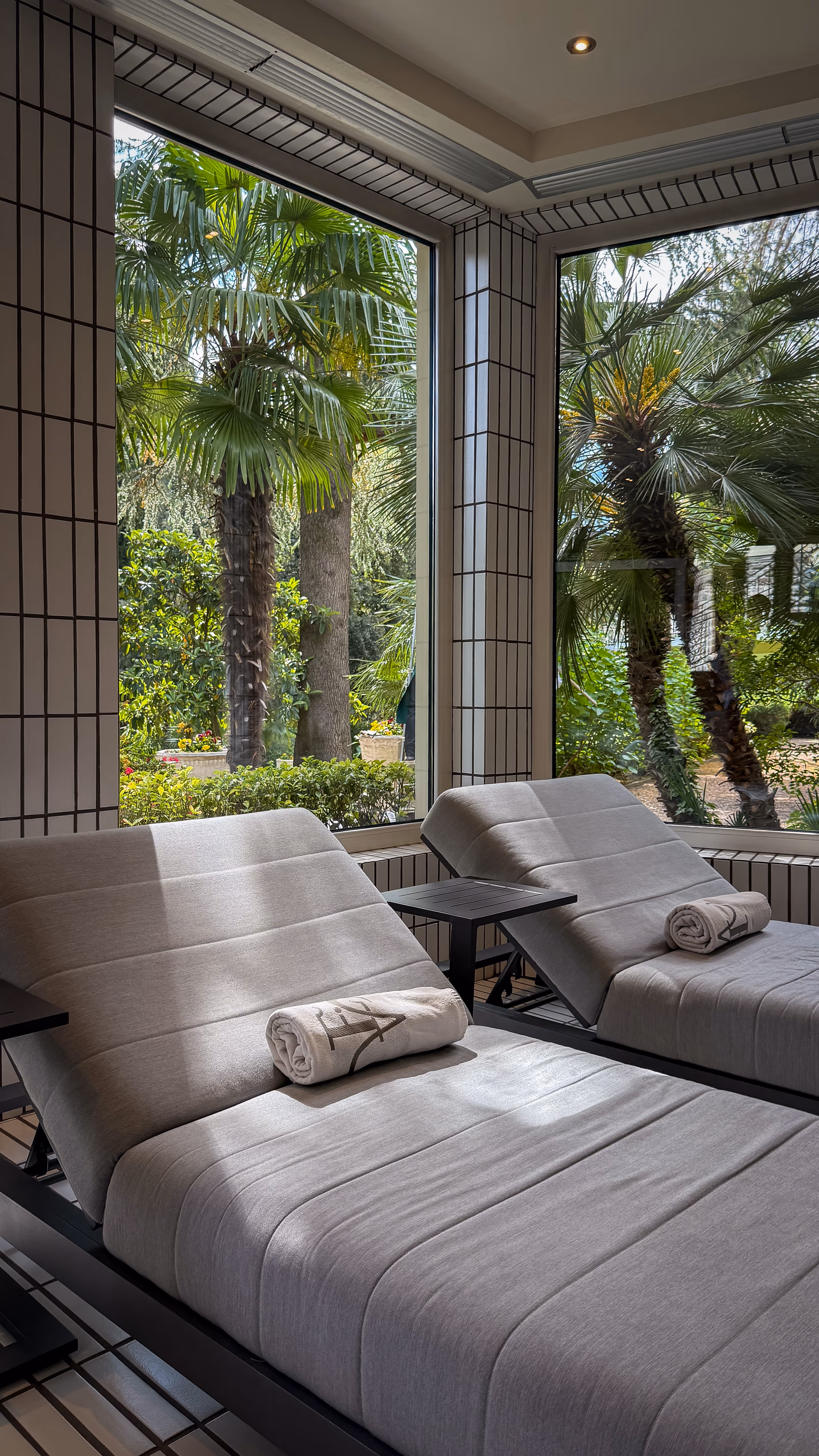 Two gray lounge chairs with rolled towels face large windows showing tropical garden with palm trees.