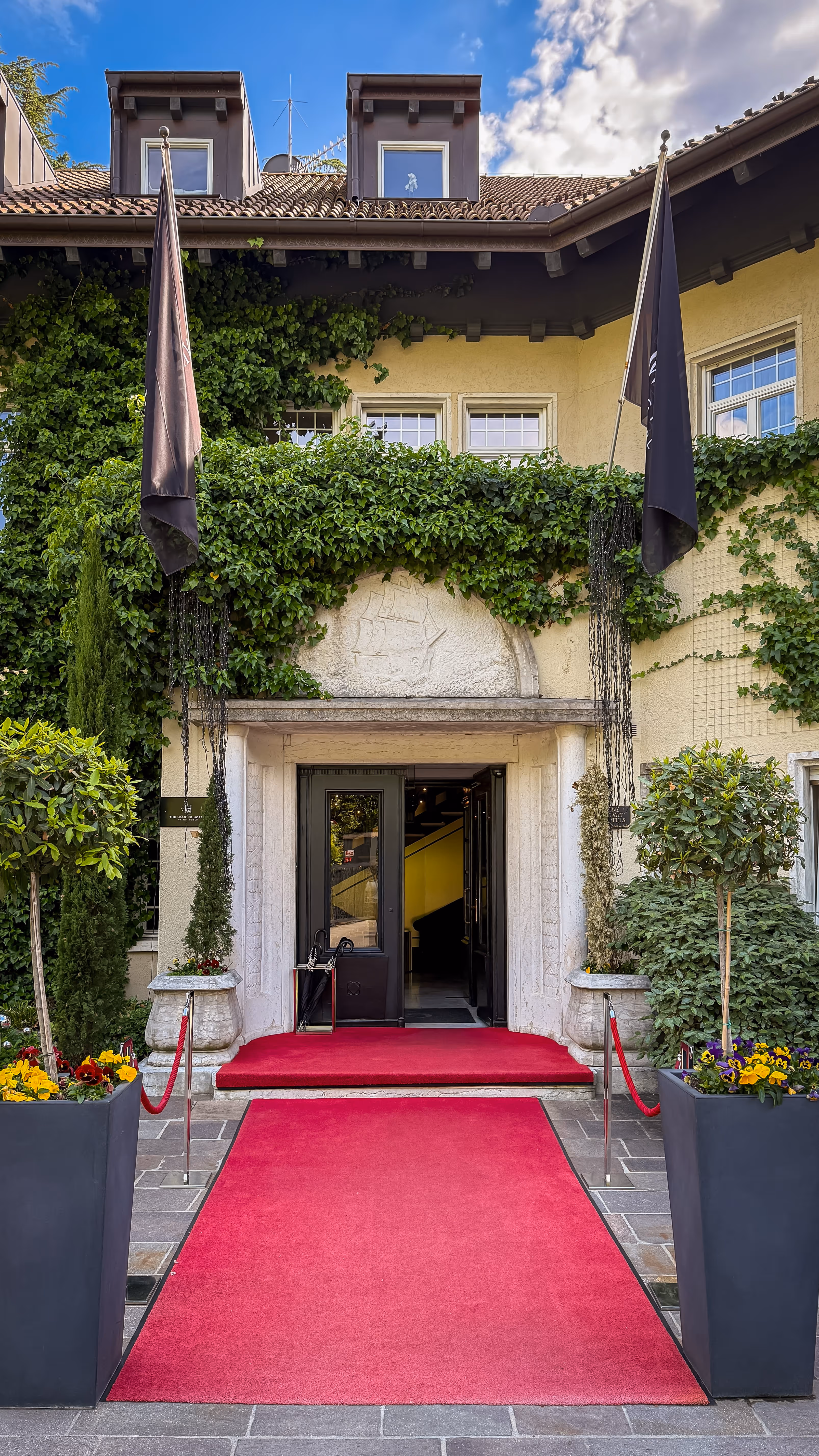 Entryway with red carpet, potted plants, ivy-covered walls, and open glass doors to a building.