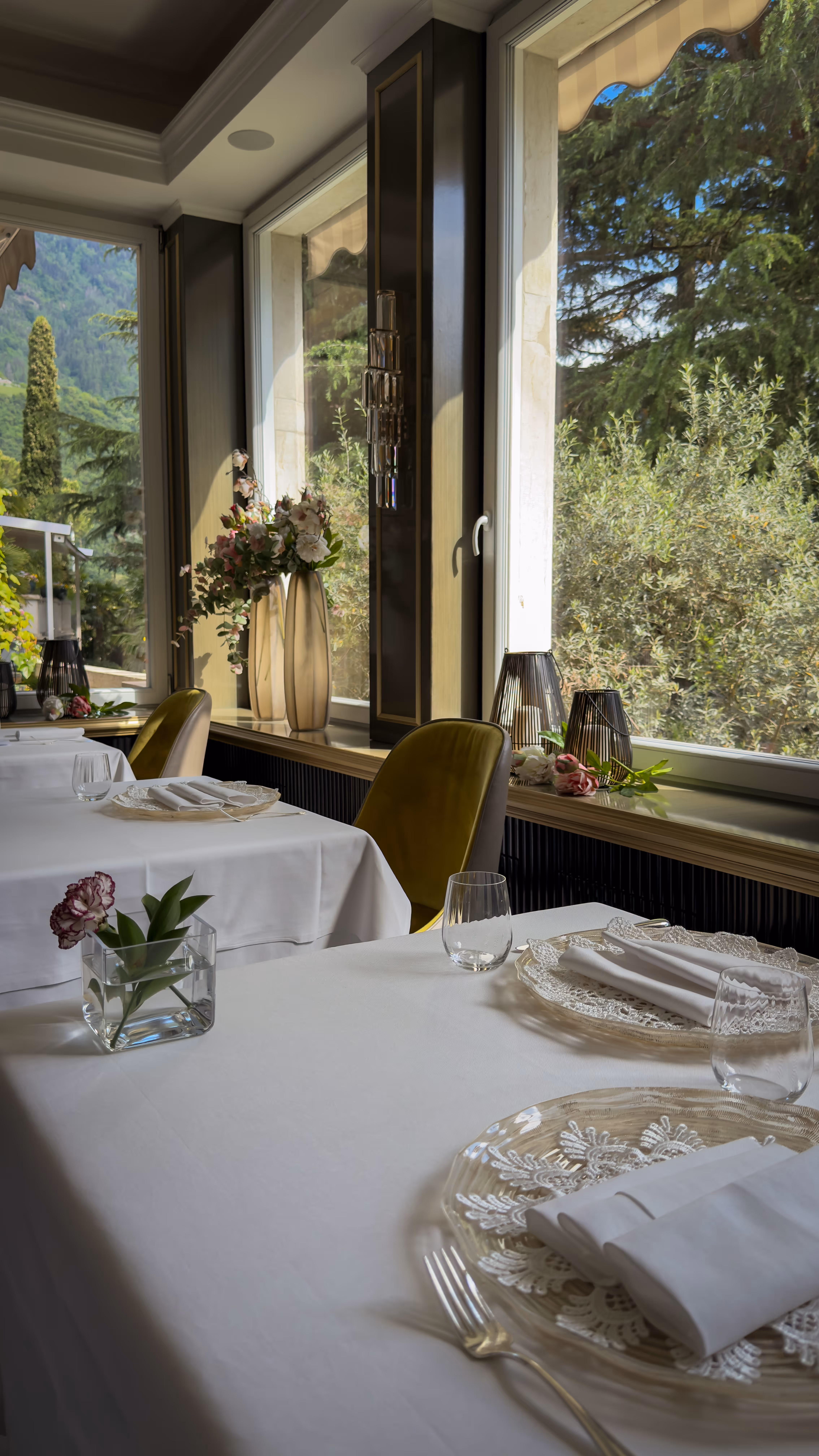 Elegant dining tables with white cloth, glass plates, folded napkins, flowers, and large windows with greenery view.