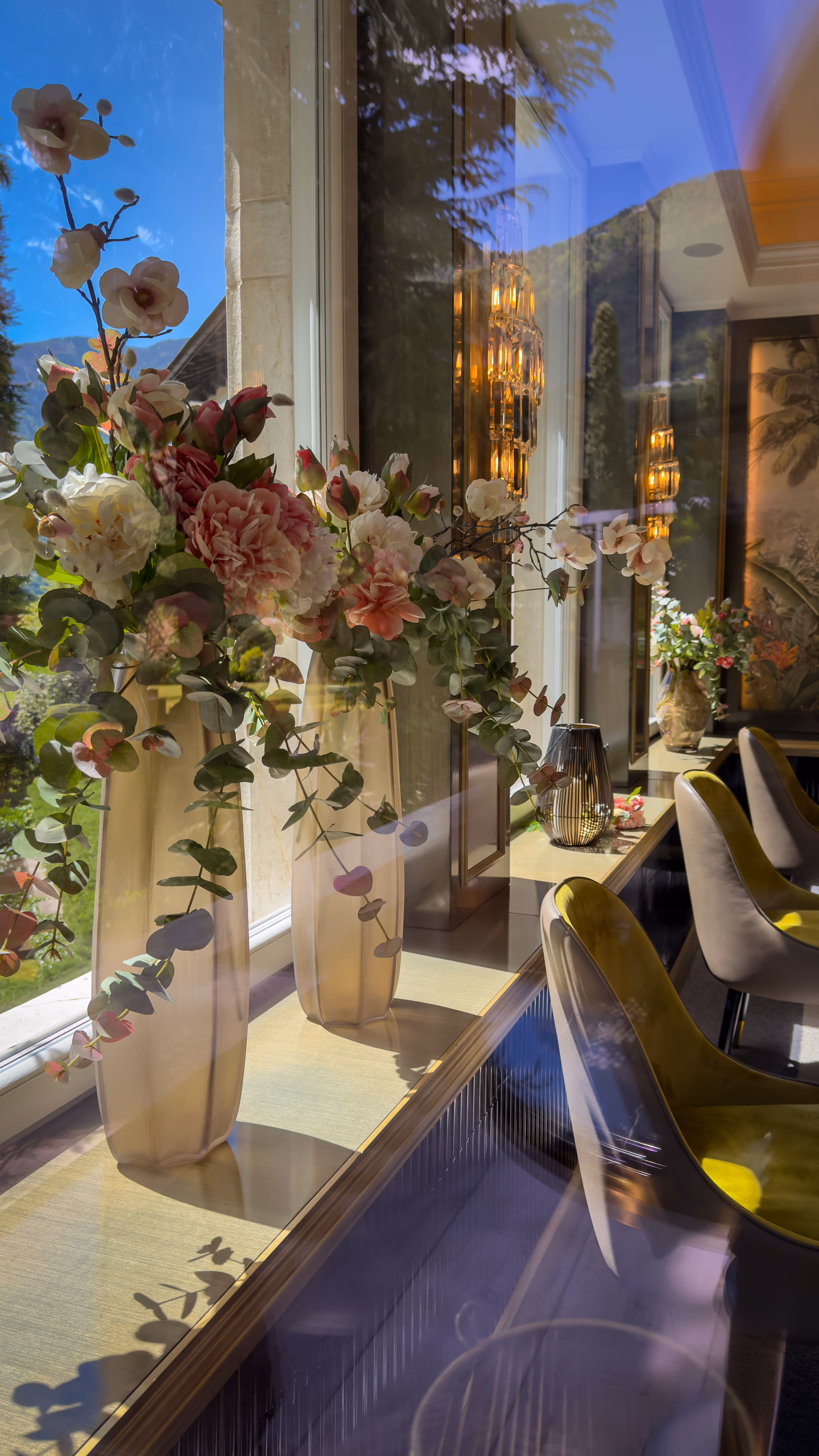 Vases with pink and white flowers on a sunny windowsill, with chairs and wall lights inside.
