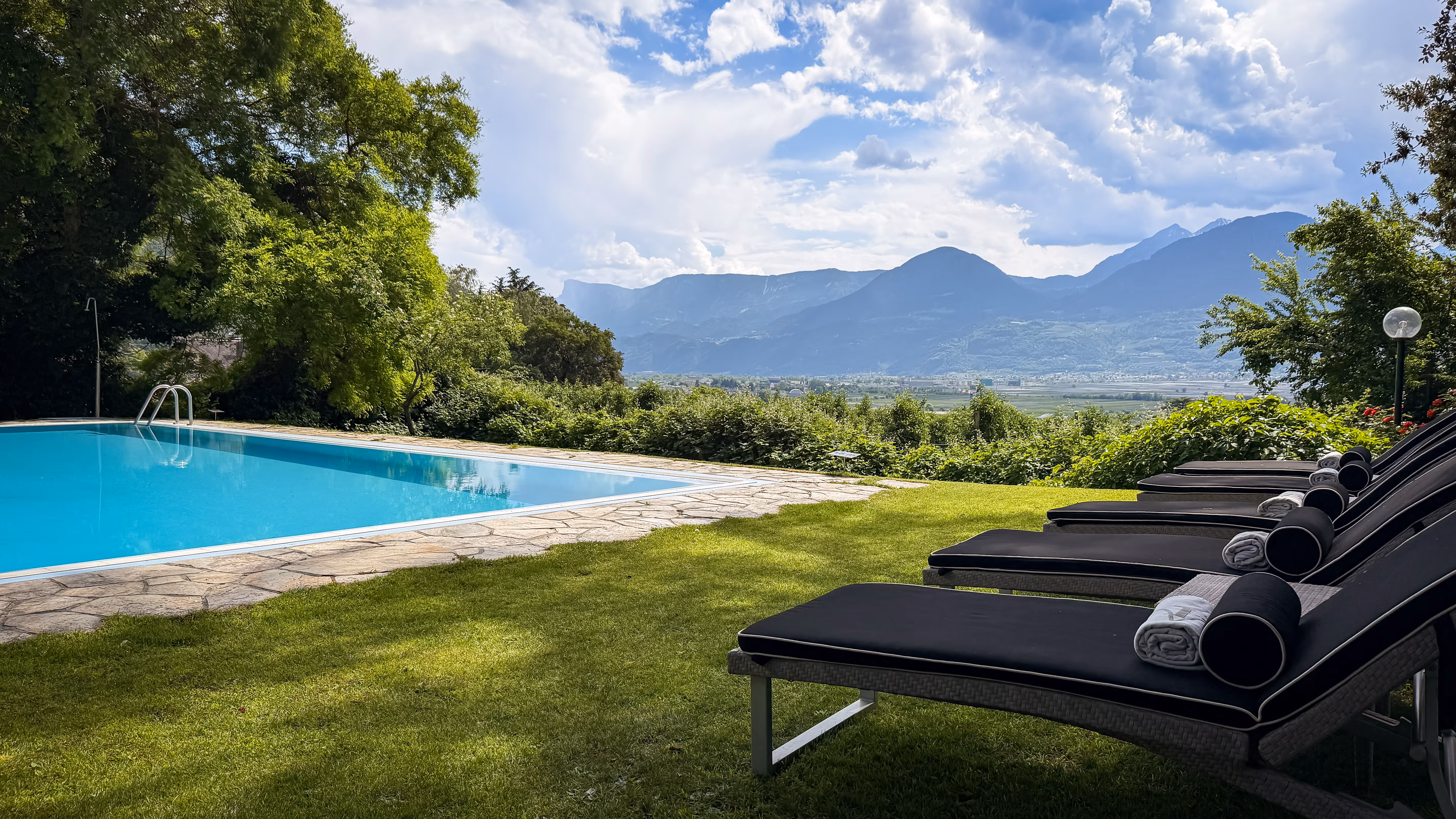 Outdoor pool beside a row of black cushioned lounge chairs on grass with mountains in the background.