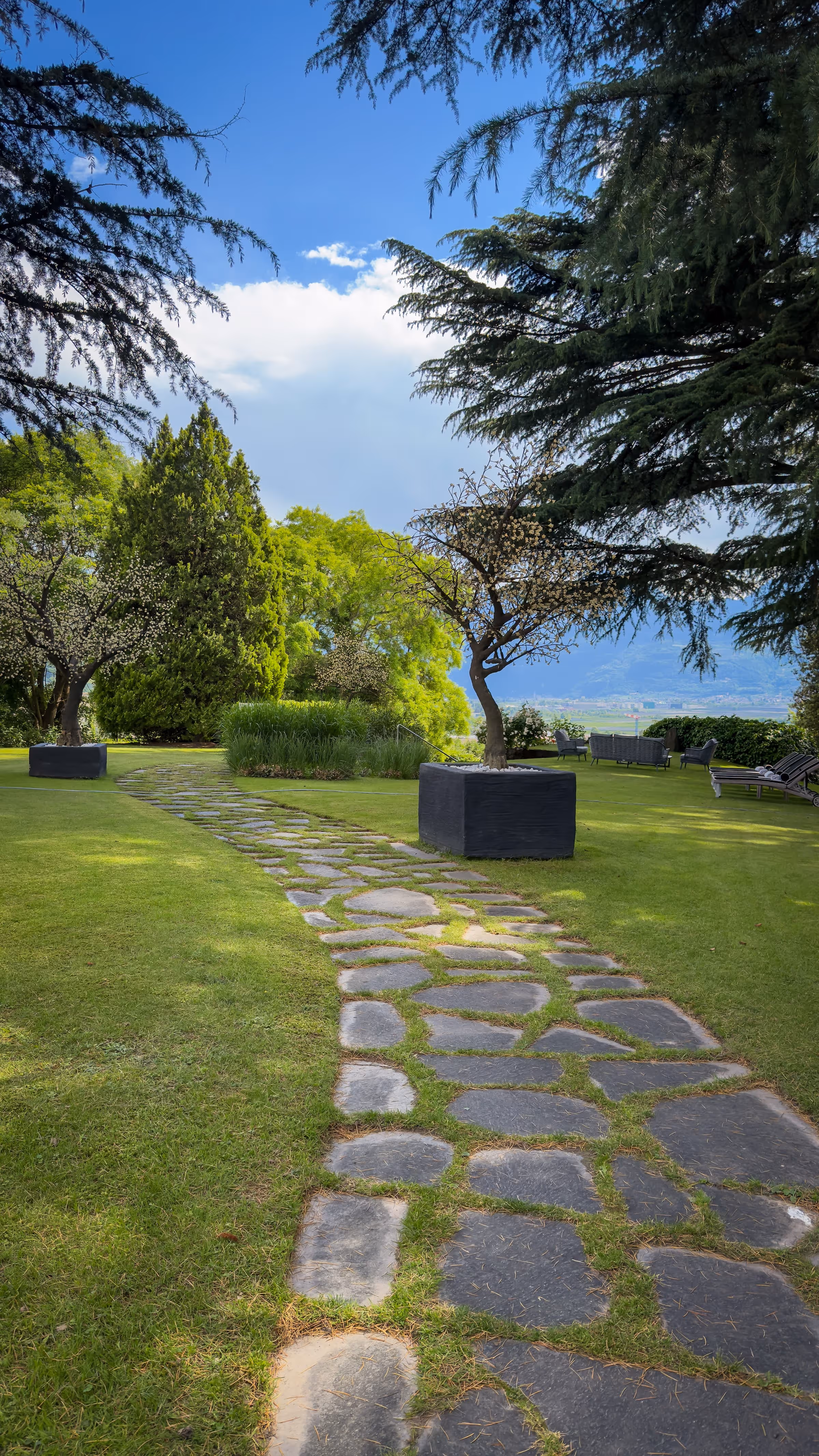 Curved stone path through lush garden with trees, green grass, and blue sky.