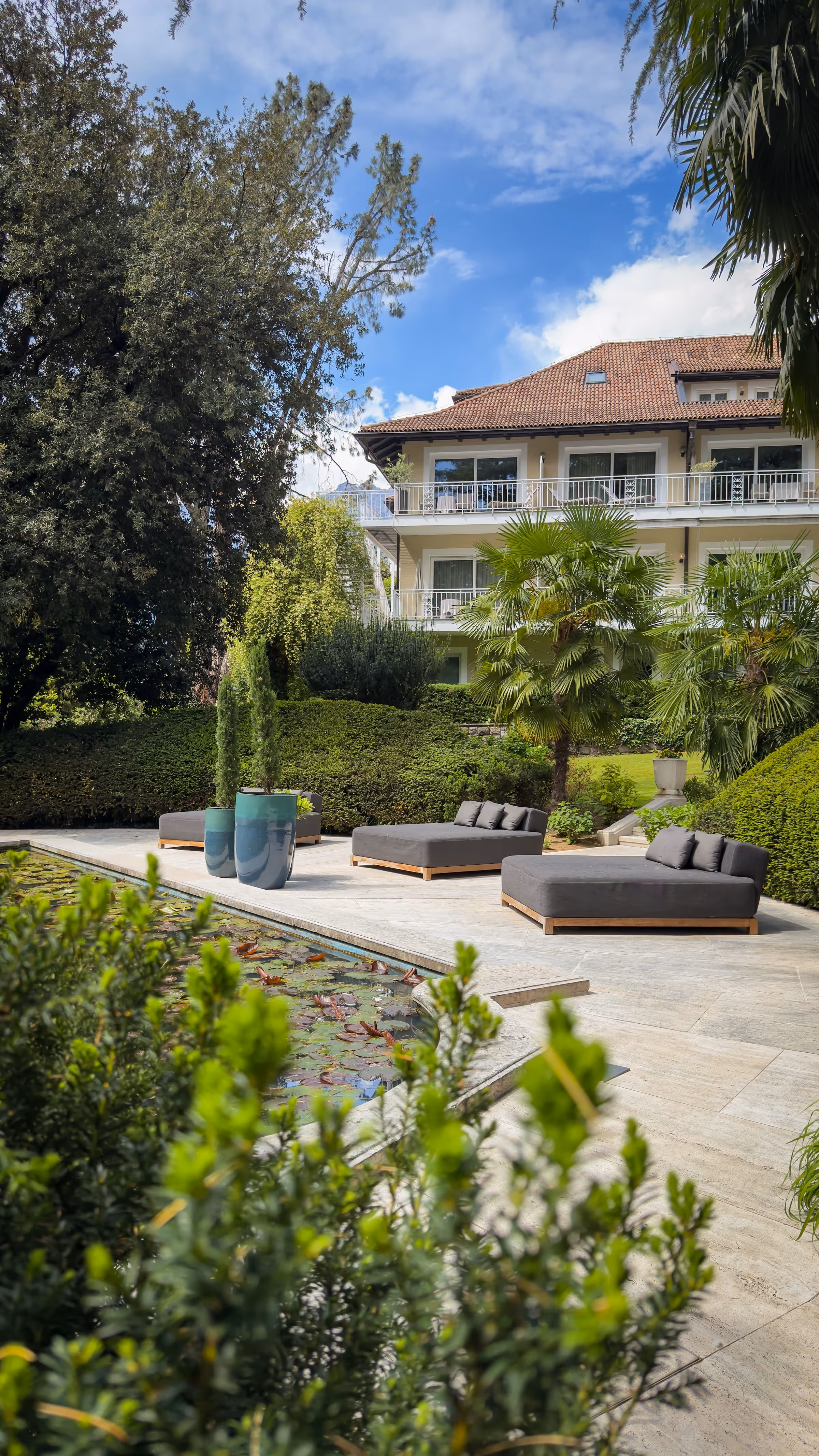 Modern outdoor patio with gray cushioned lounge chairs, plants, and a water pool in front of a house.