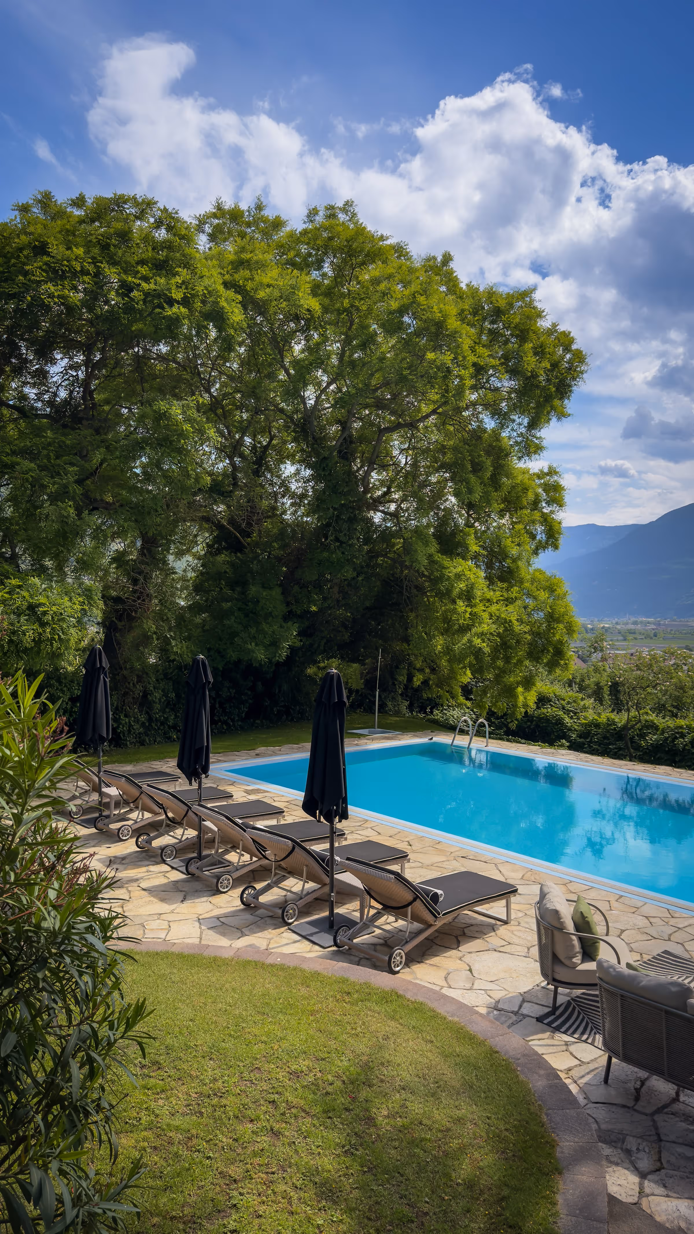Outdoor pool with blue water, six black lounge chairs with closed umbrellas, trees and mountains in background.