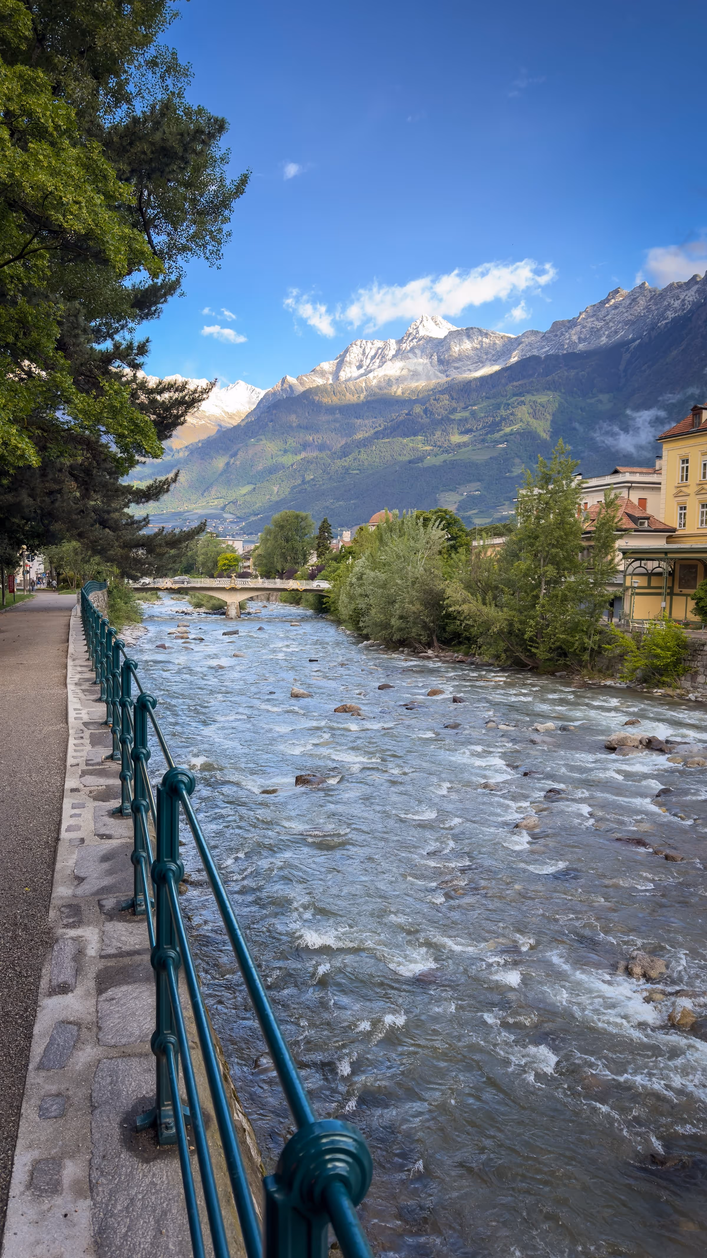 Clear river flowing beside a tree-lined path with snowy mountains and buildings in the background.