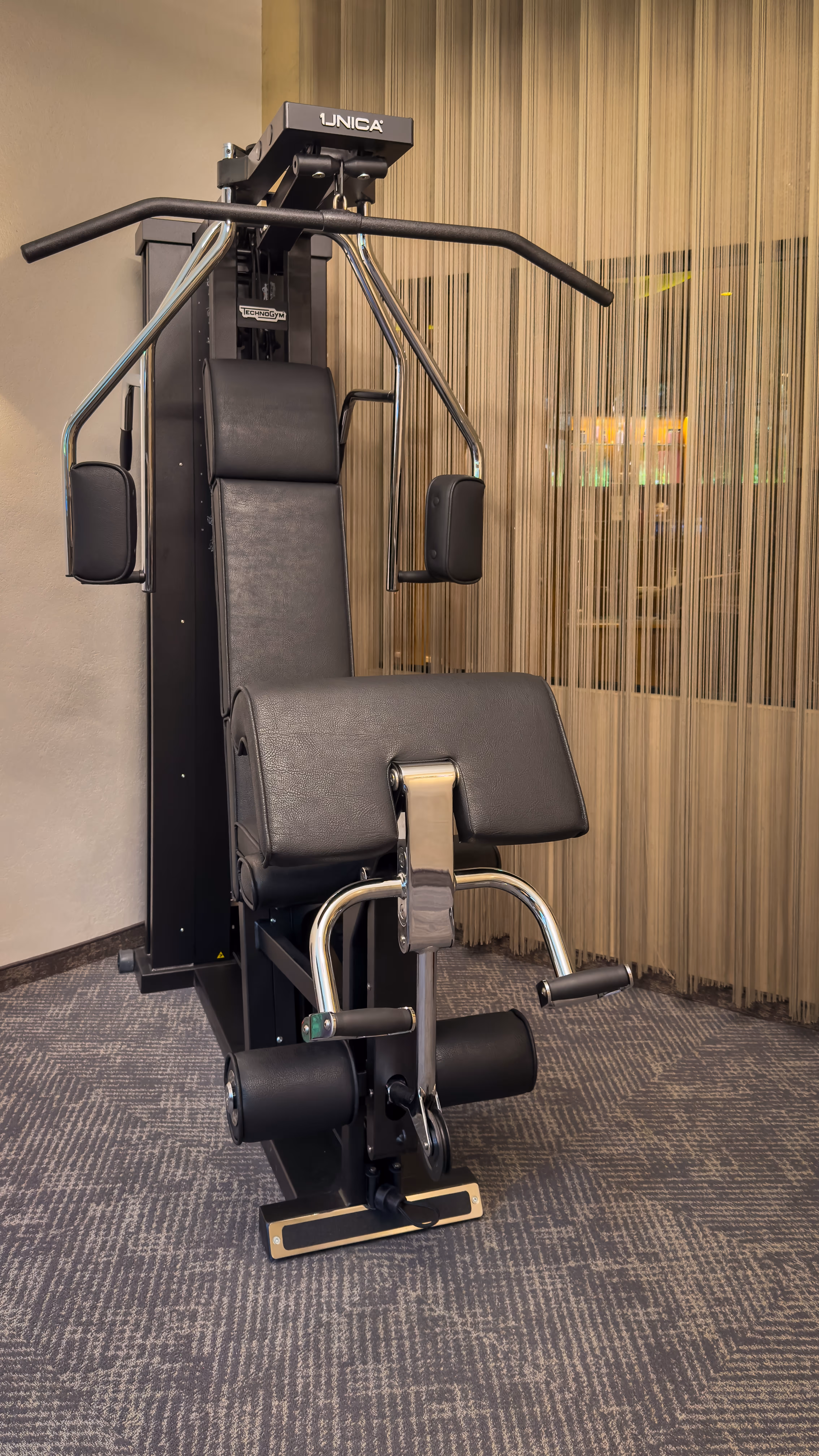 Black Technogym Uniqa gym machine for strength training in a room with patterned carpeting.