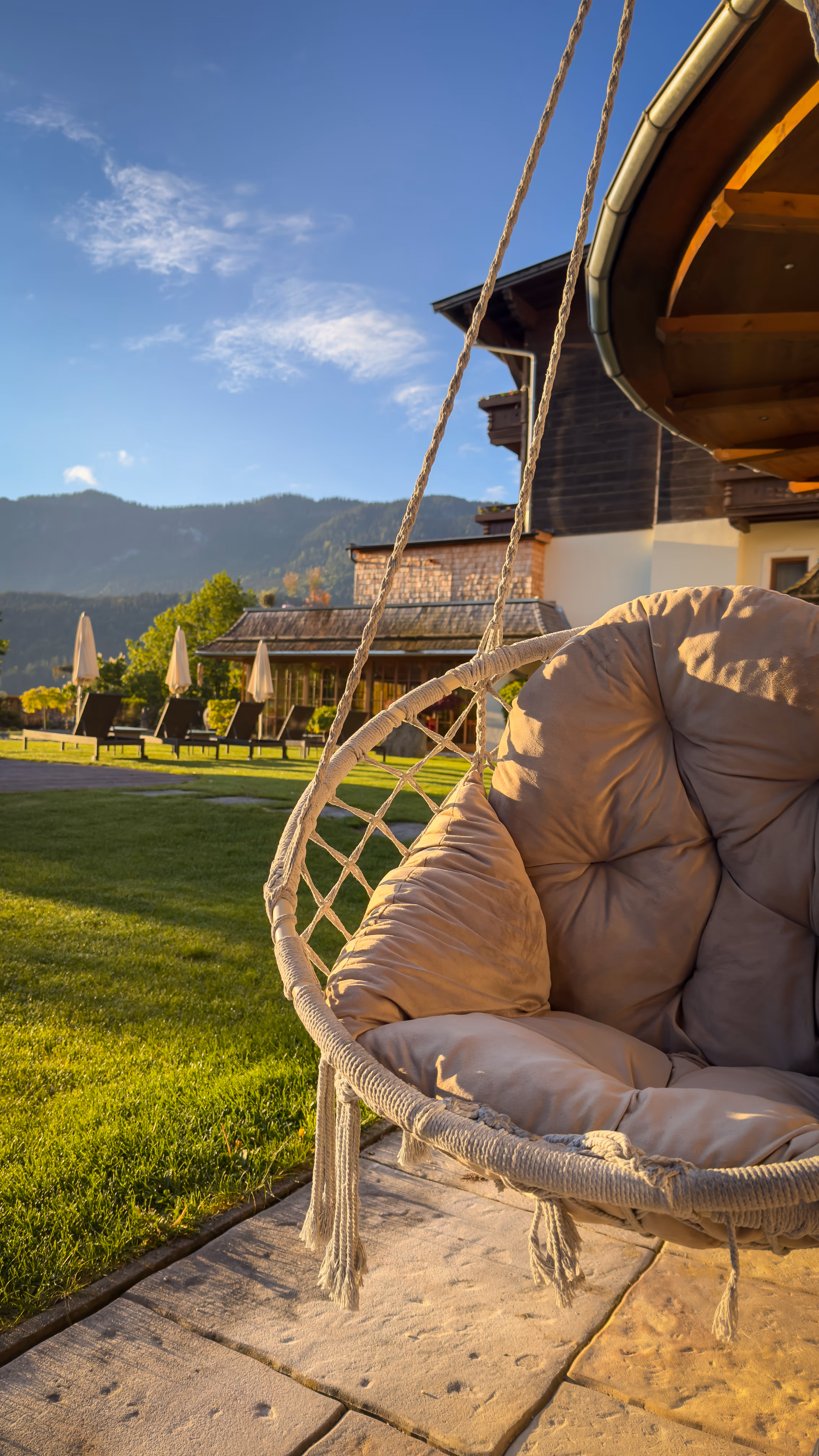 Cozy cushioned hanging chair outdoors with green lawn, umbrellas, and mountains in the background.
