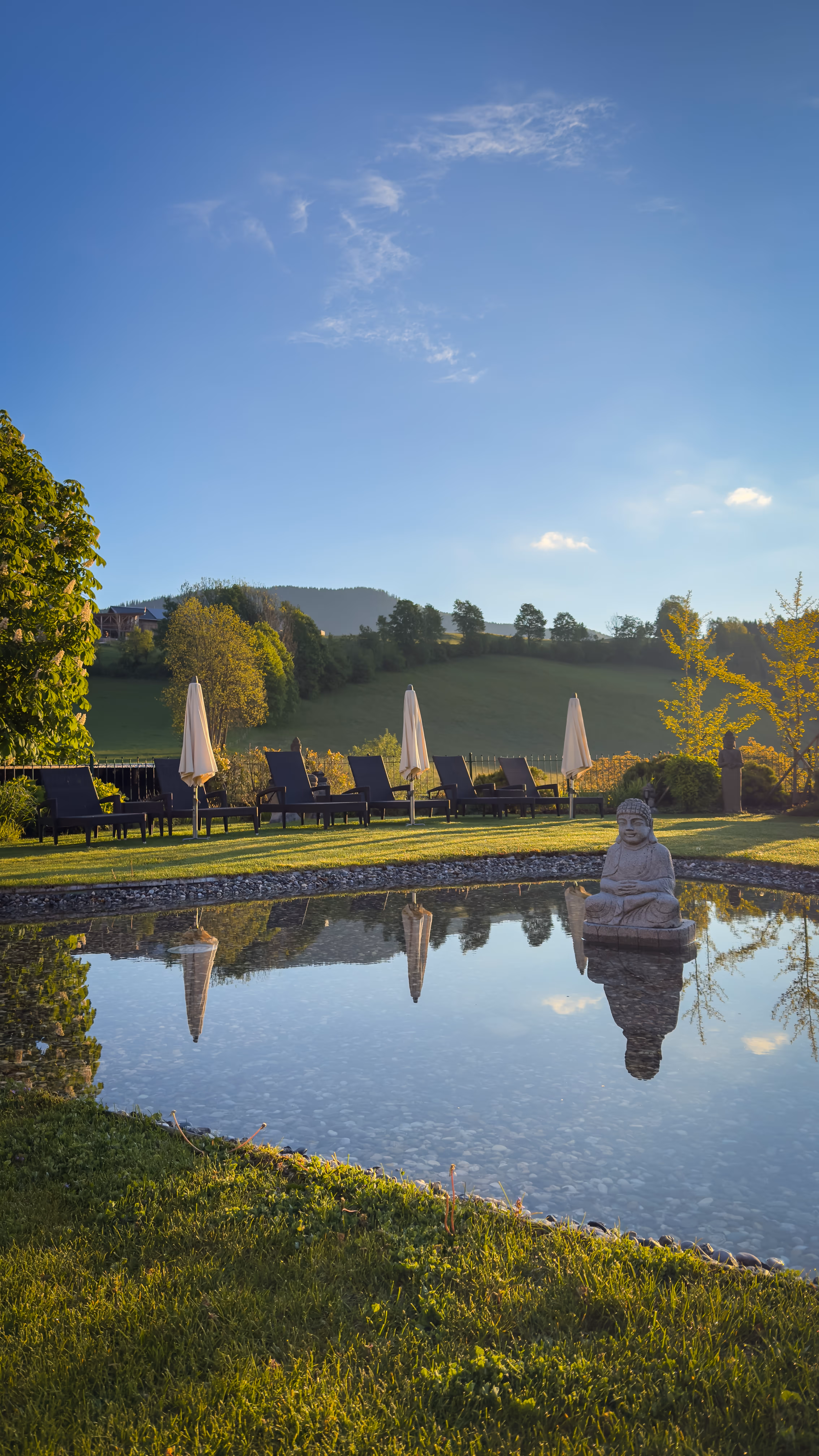 Calm garden pond with a Buddha statue reflecting in water, lawn chairs and umbrellas on grass.