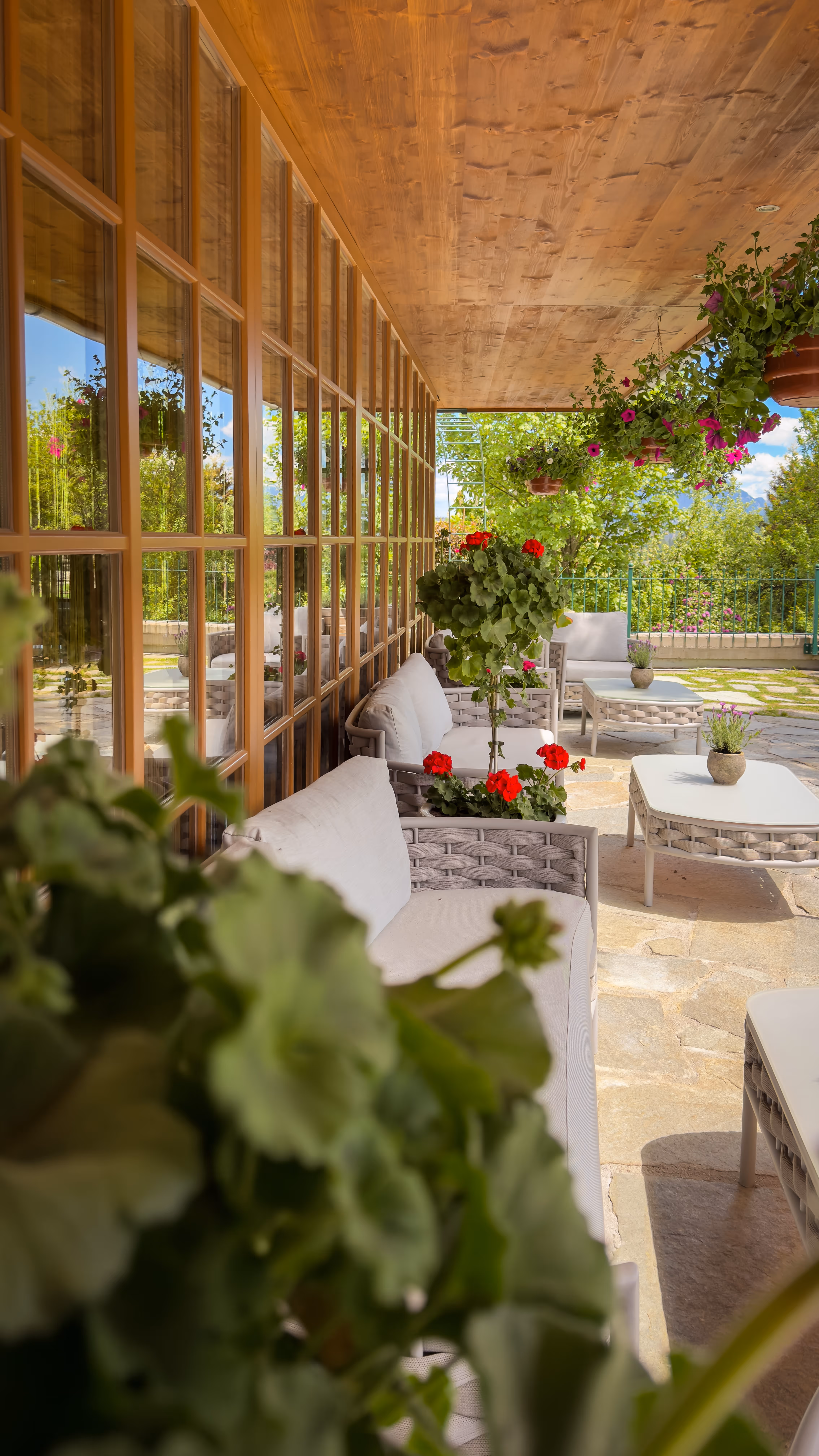 Outdoor patio with wicker sofas, white cushions, hanging plants, and red flowers under a wooden roof.