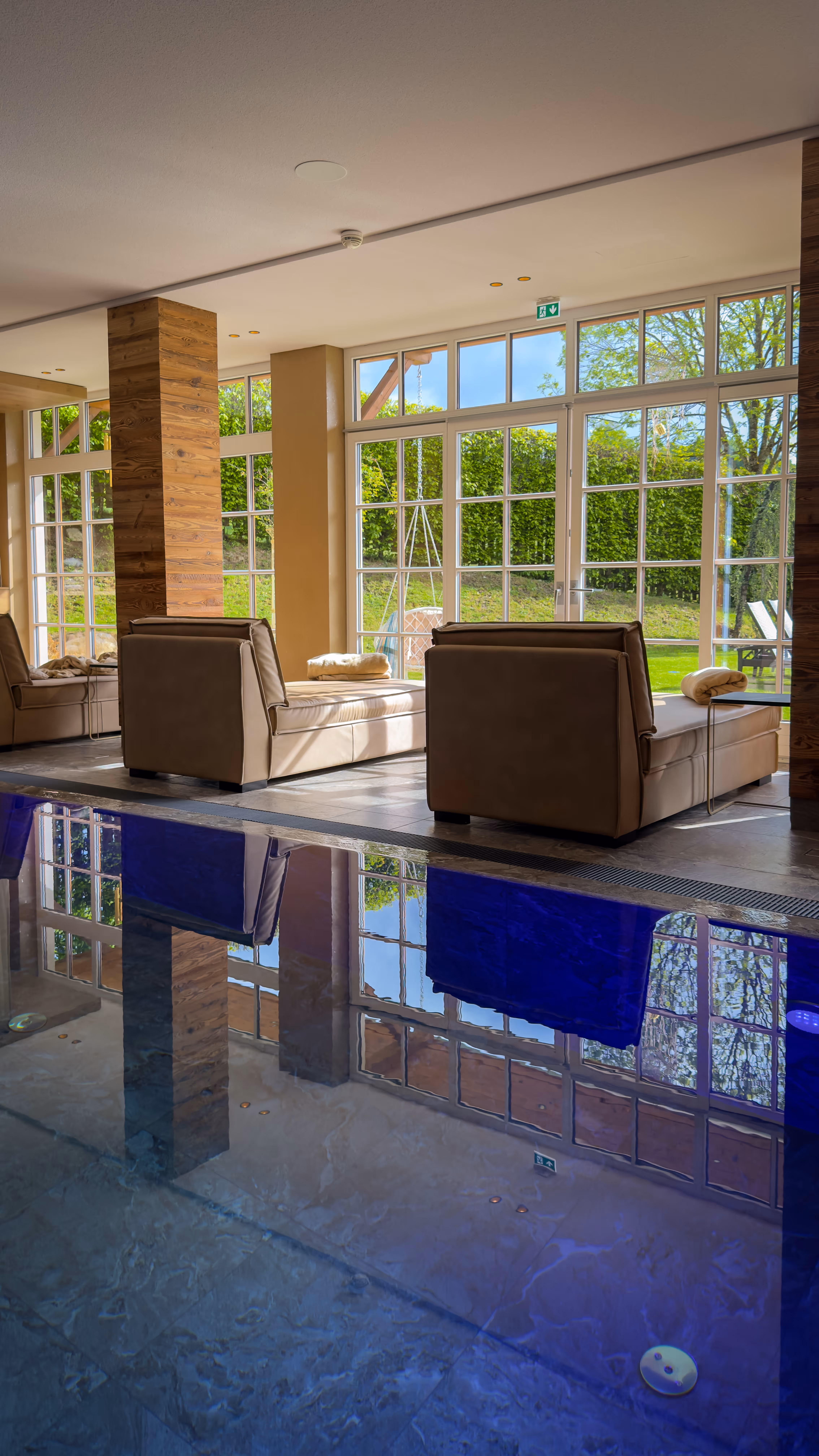 Indoor pool area with beige lounge chairs near large windows showing green outdoor garden.