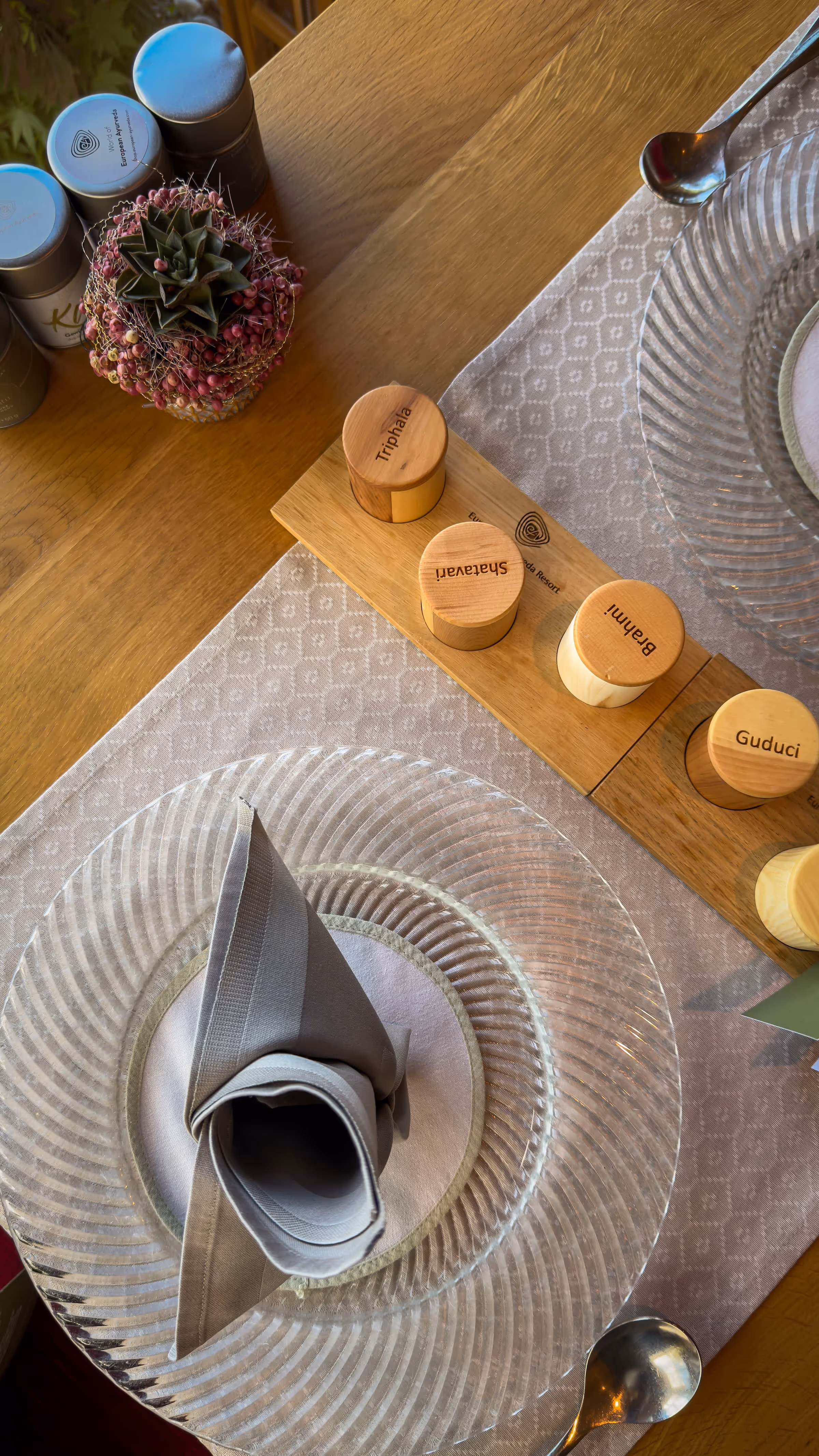 Elegant table setting with glass plate, folded gray napkin, wooden spice containers, and succulent plant.