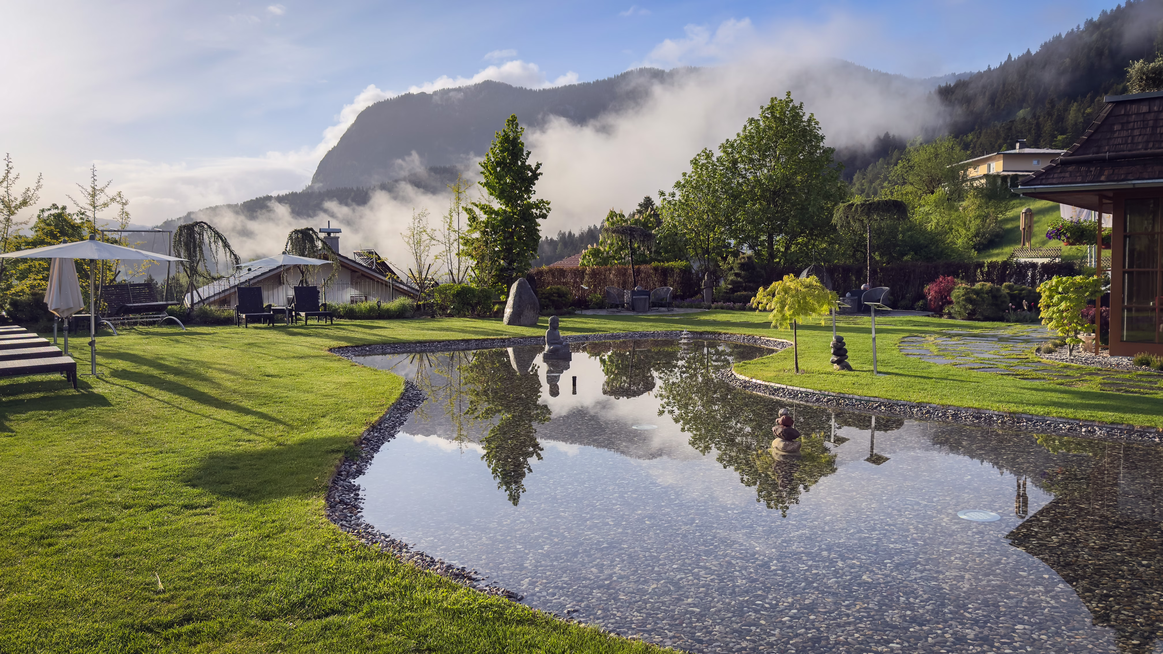 Outdoor pool with sun loungers, red flowers, green lawn, trees, and forested mountains in background.