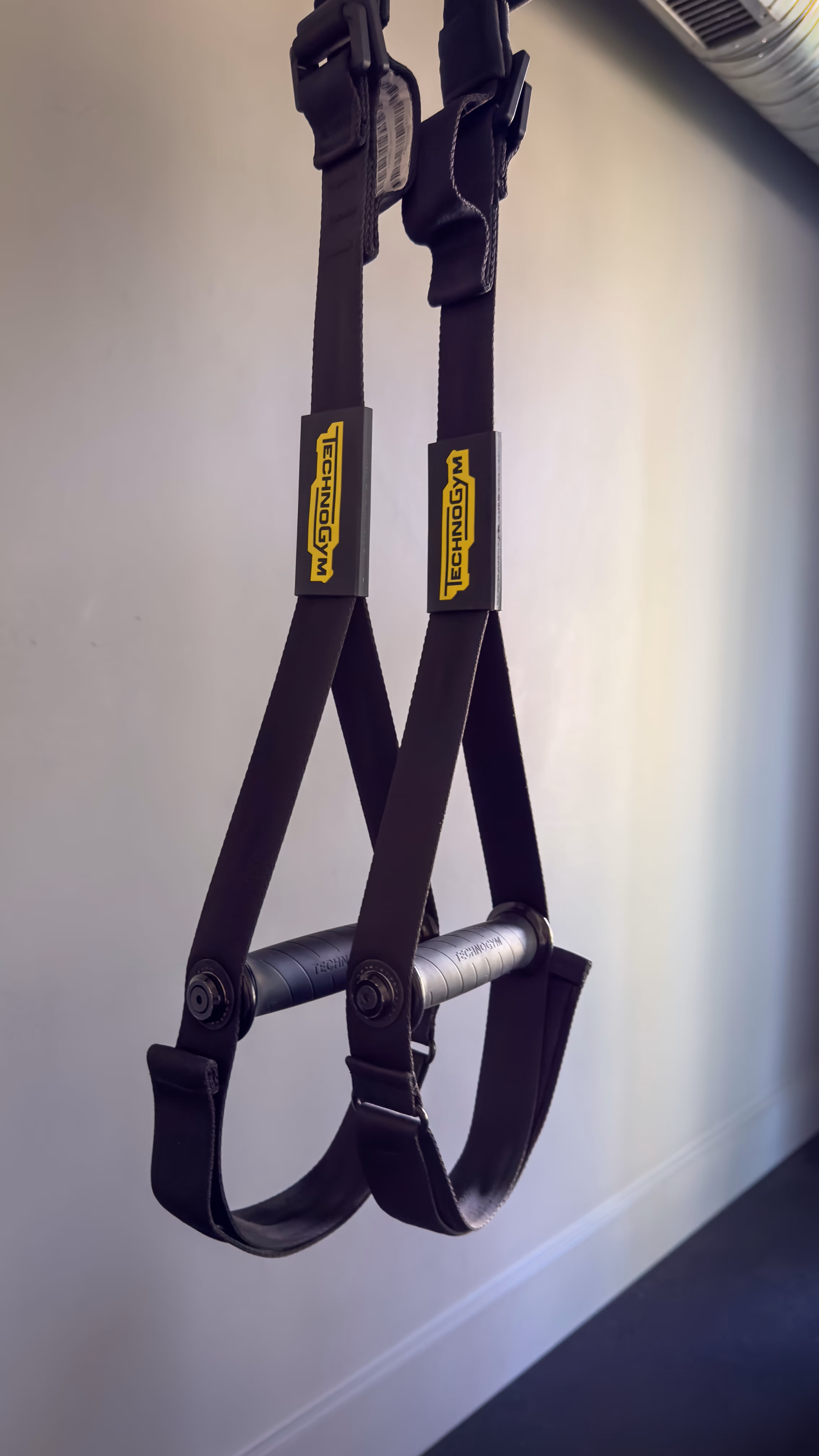 Black Technogym suspension training straps with handles hanging indoors against a white wall.