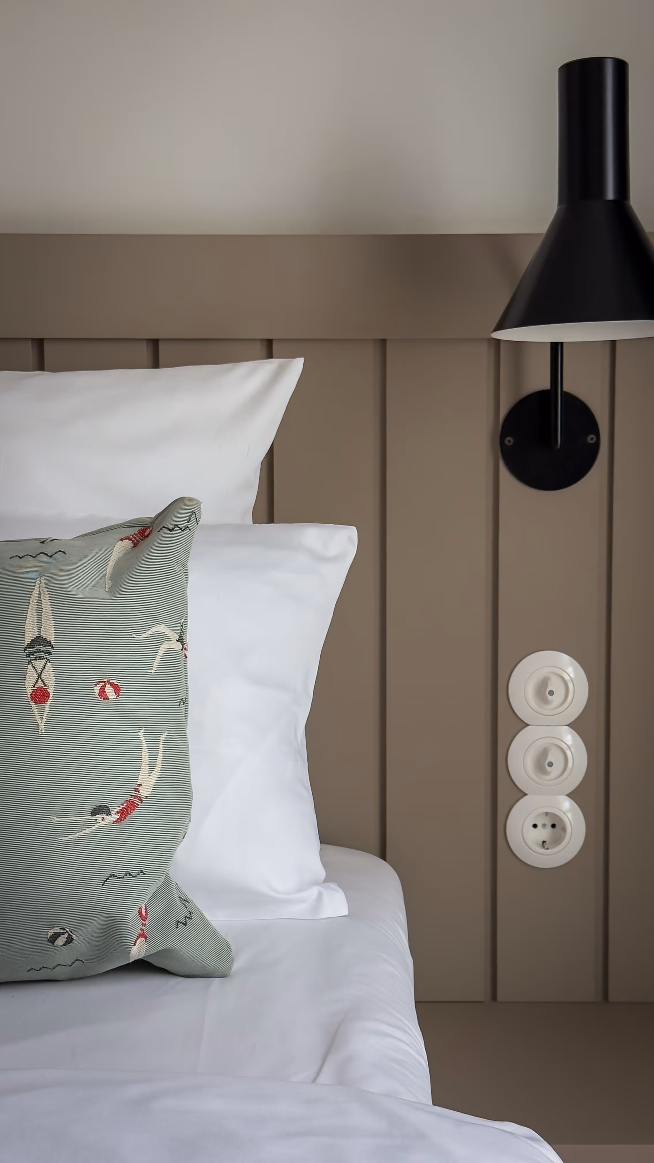 Bed with white pillows and a decorative pillow with swimmers, beside a black wall lamp and three white switches.