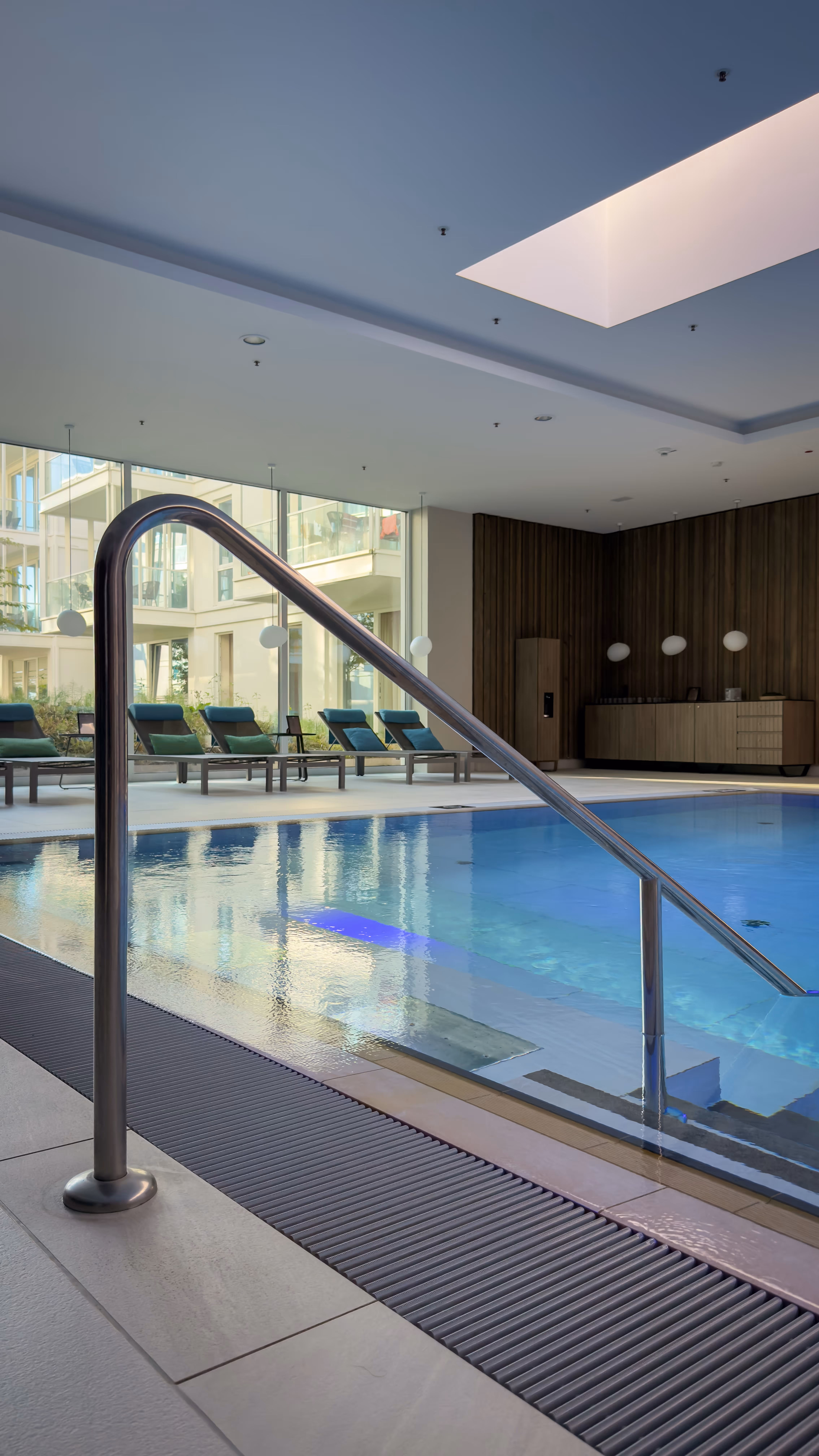 Indoor swimming pool with metal handrail, lounge chairs, and large windows showing building exterior.