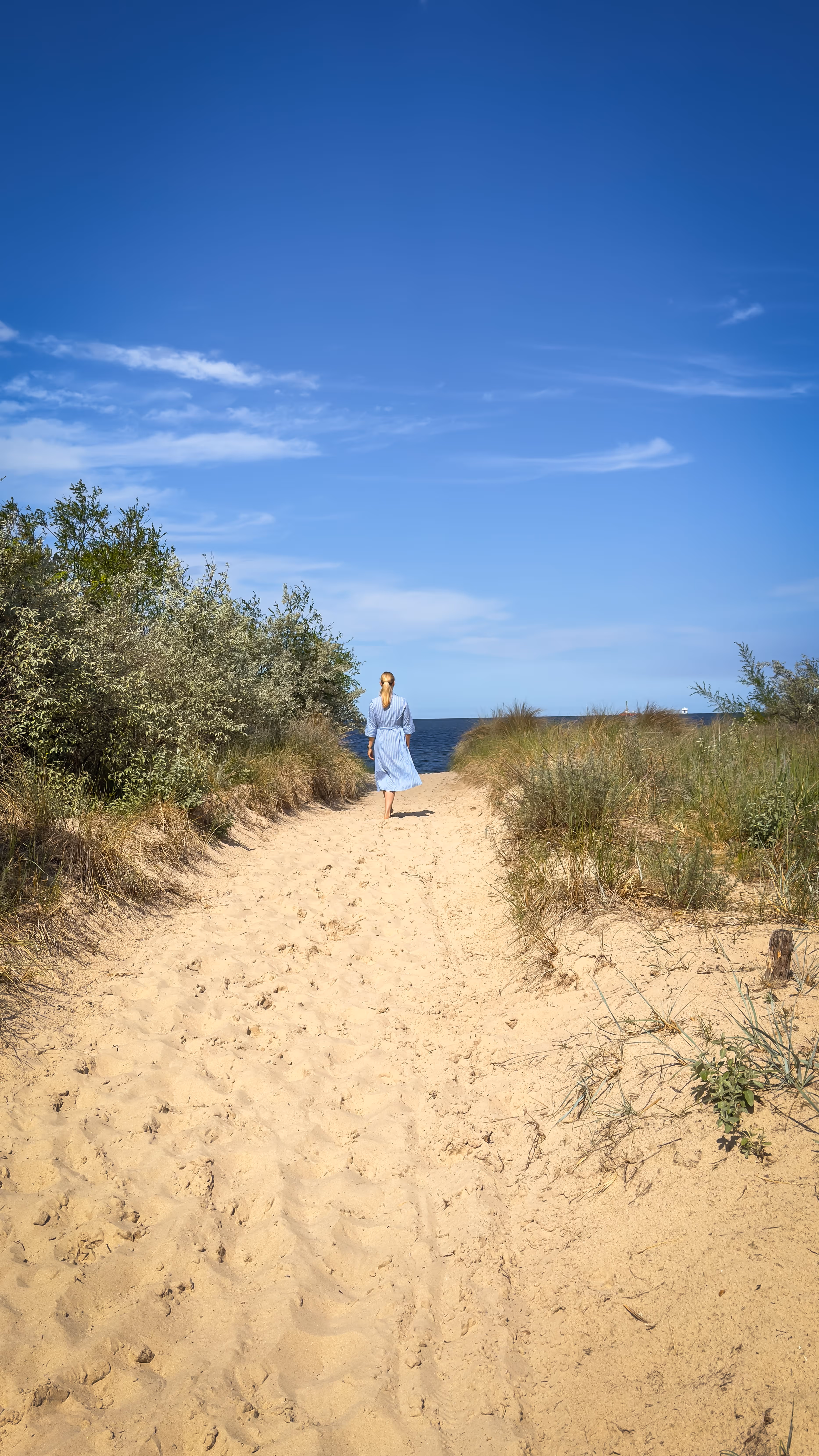 Woman in blue dress walking on sandy path toward the ocean under a clear blue sky.