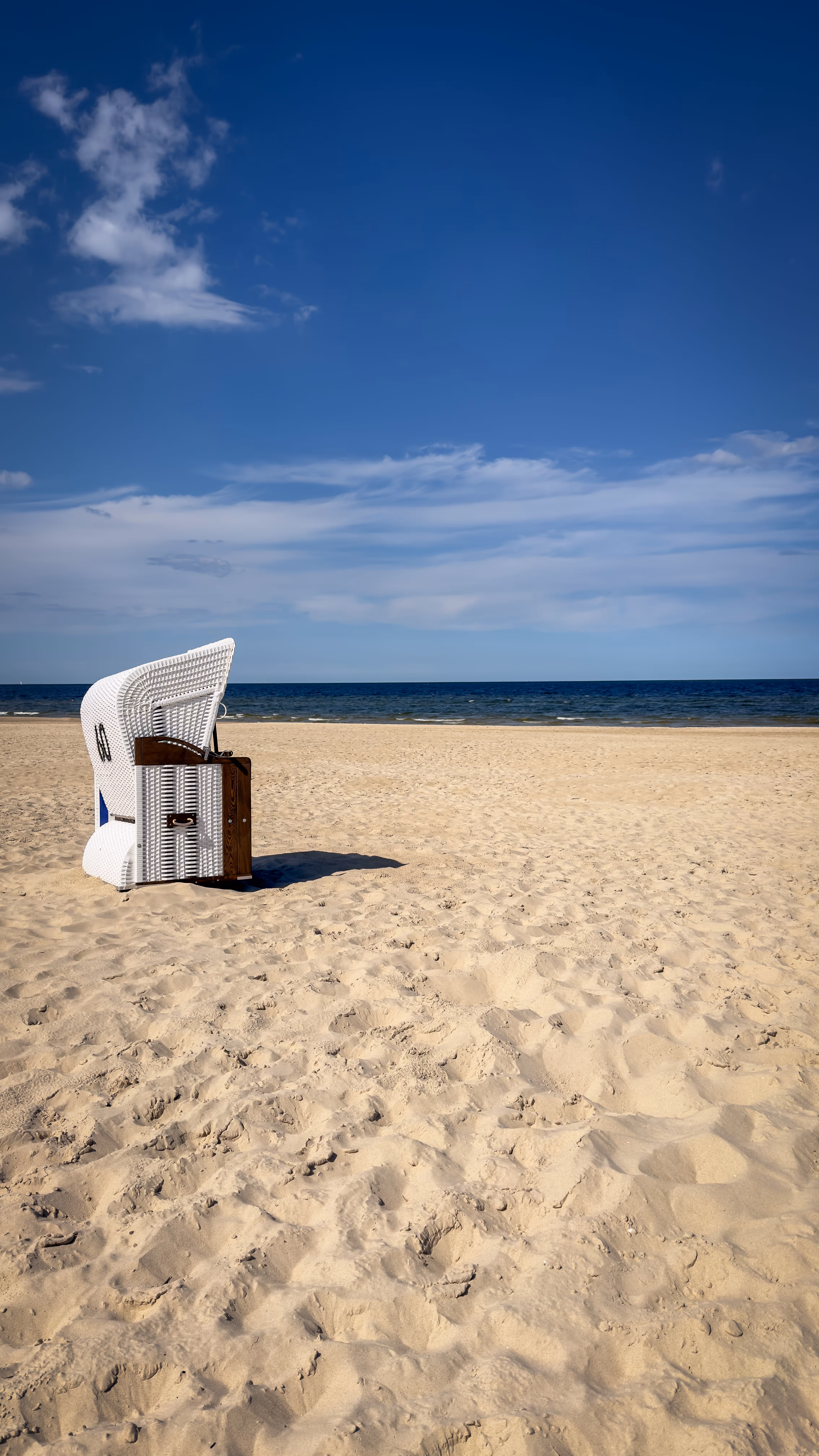 Empty white wicker beach chair on sandy beach with ocean and blue sky in the background.