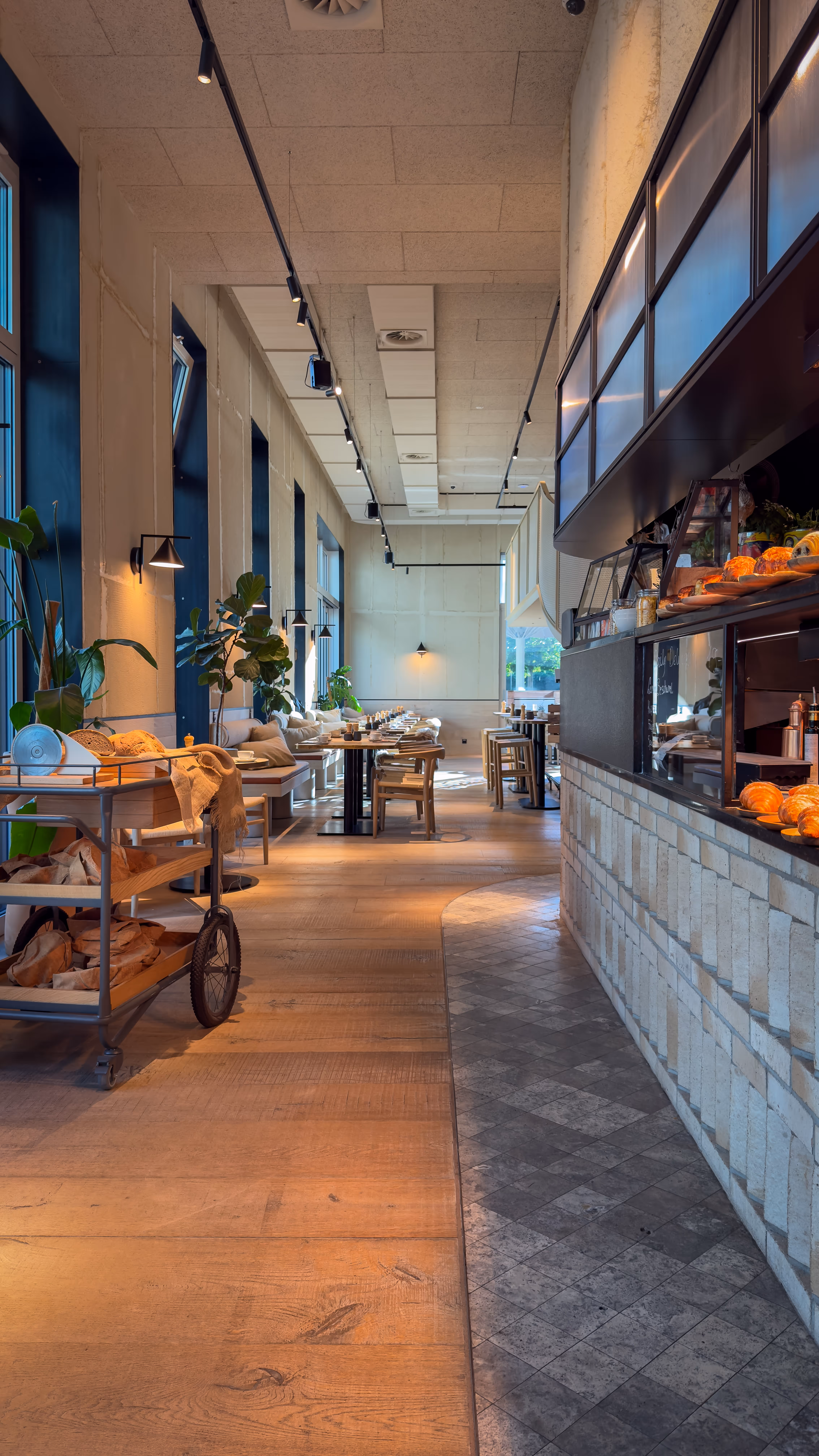 Bright modern cafe interior with tables, chairs, plants, and bread on a rolling cart.