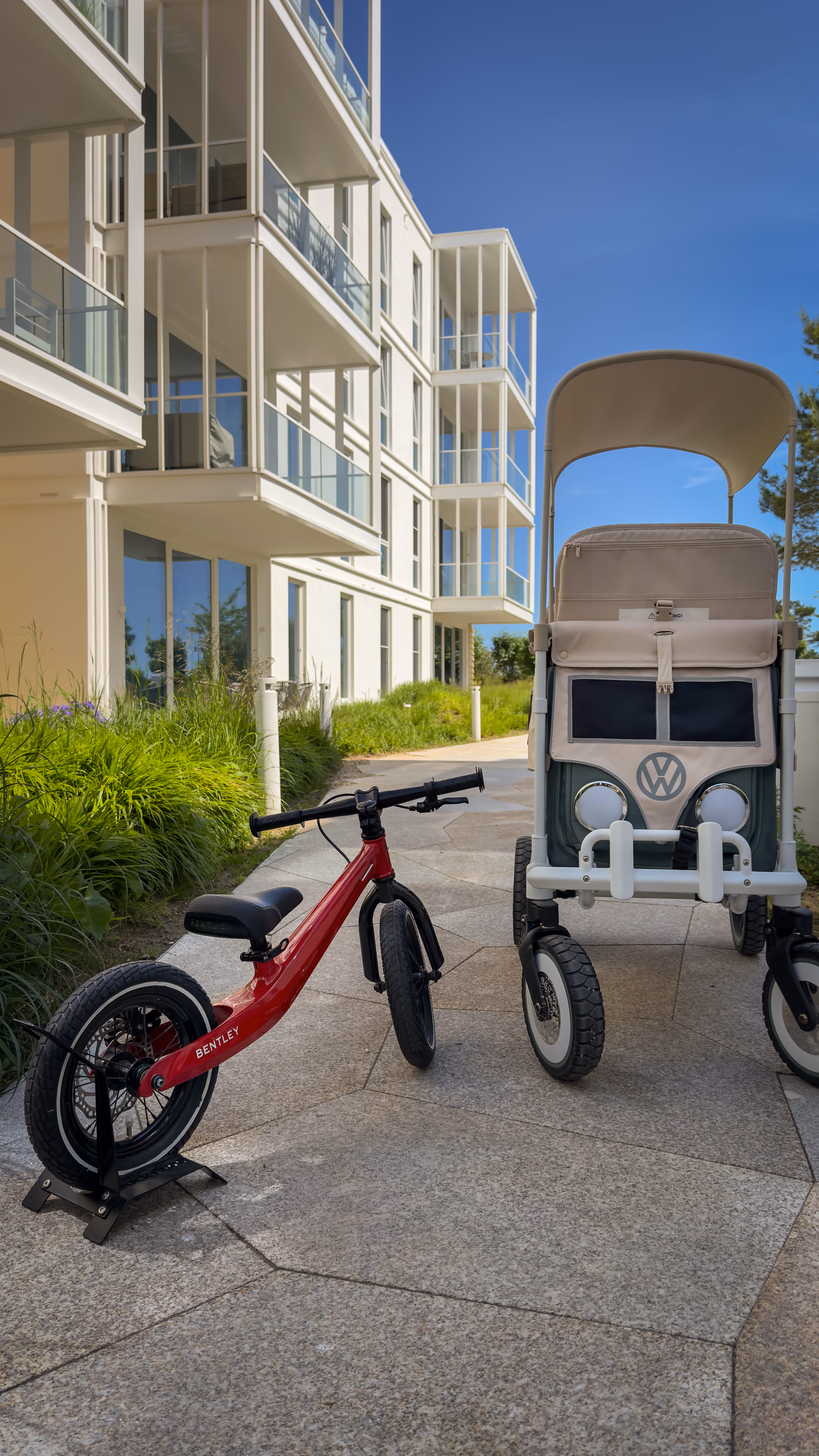 Red Bentley balance bike and beige stroller with VW logo on sidewalk near modern apartment building.