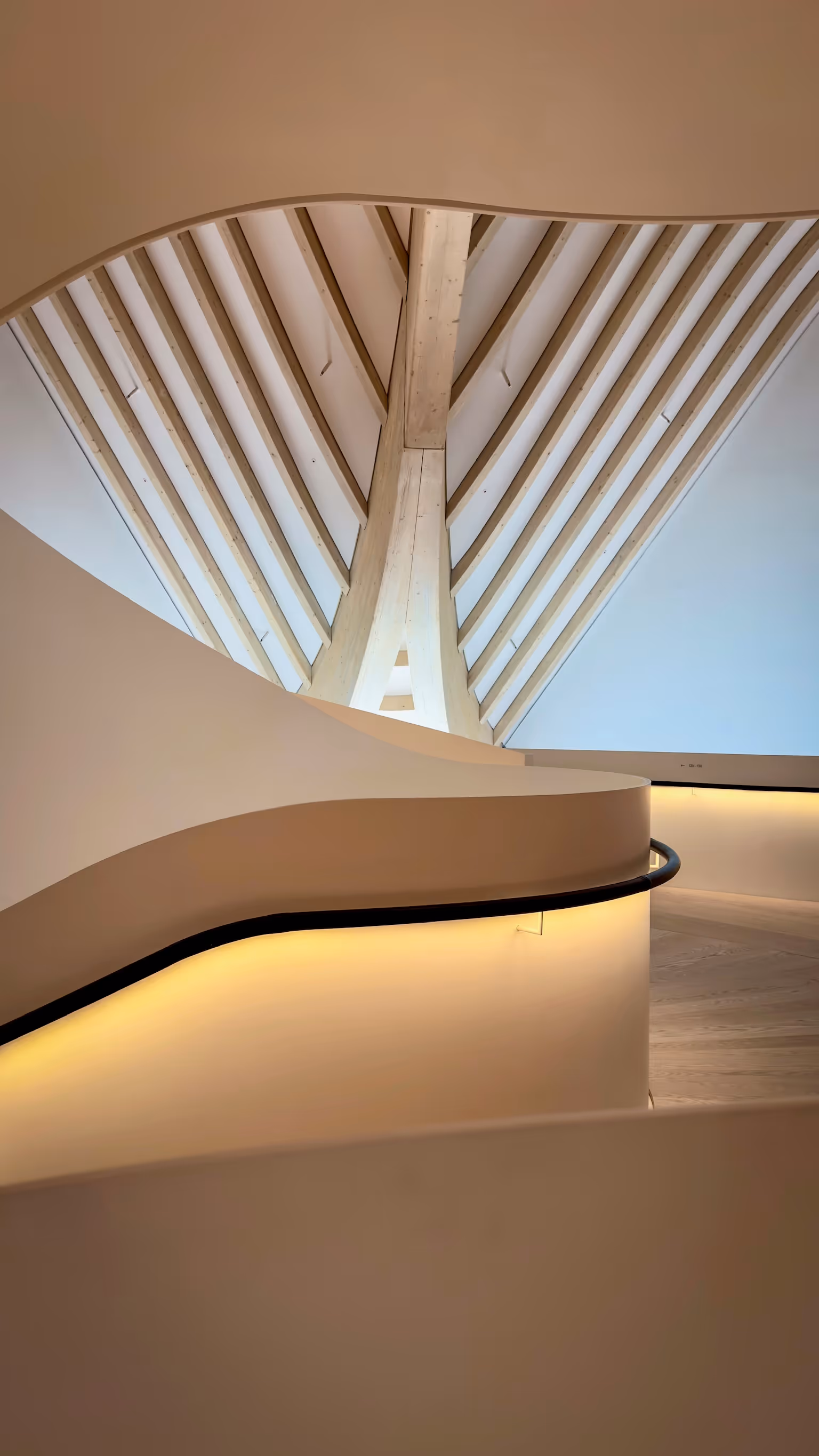 Curved, illuminated beige staircase with black handrail and wooden slatted ceiling above.