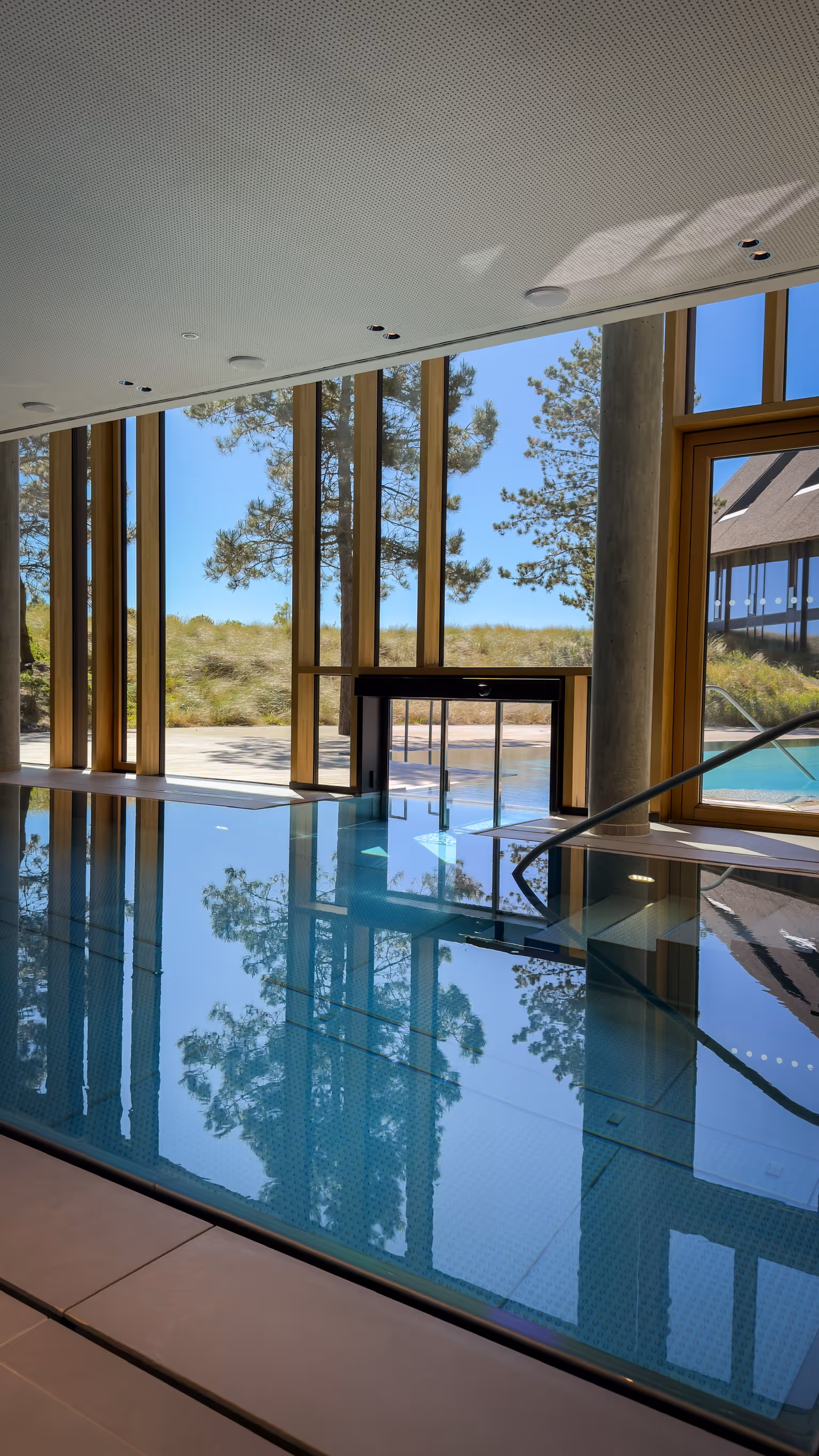 Indoor pool with glass walls reflecting trees and a view of the outdoor area with trees and blue sky.