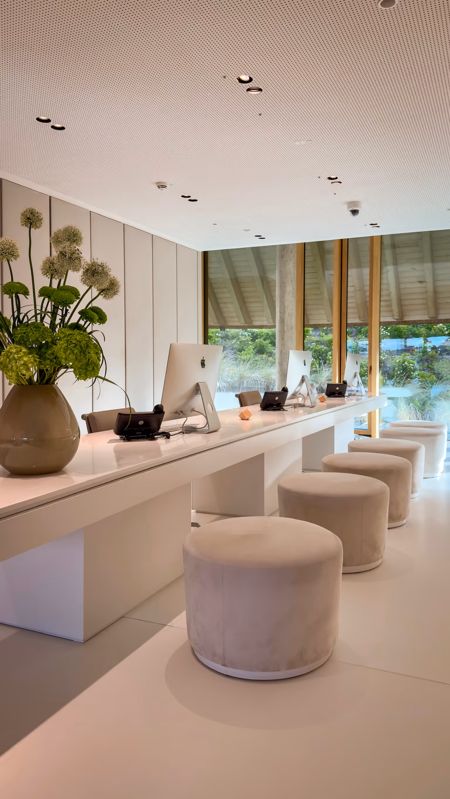 Modern office workspace with multiple iMacs, round stools, and a vase of green flowers on a long white desk.