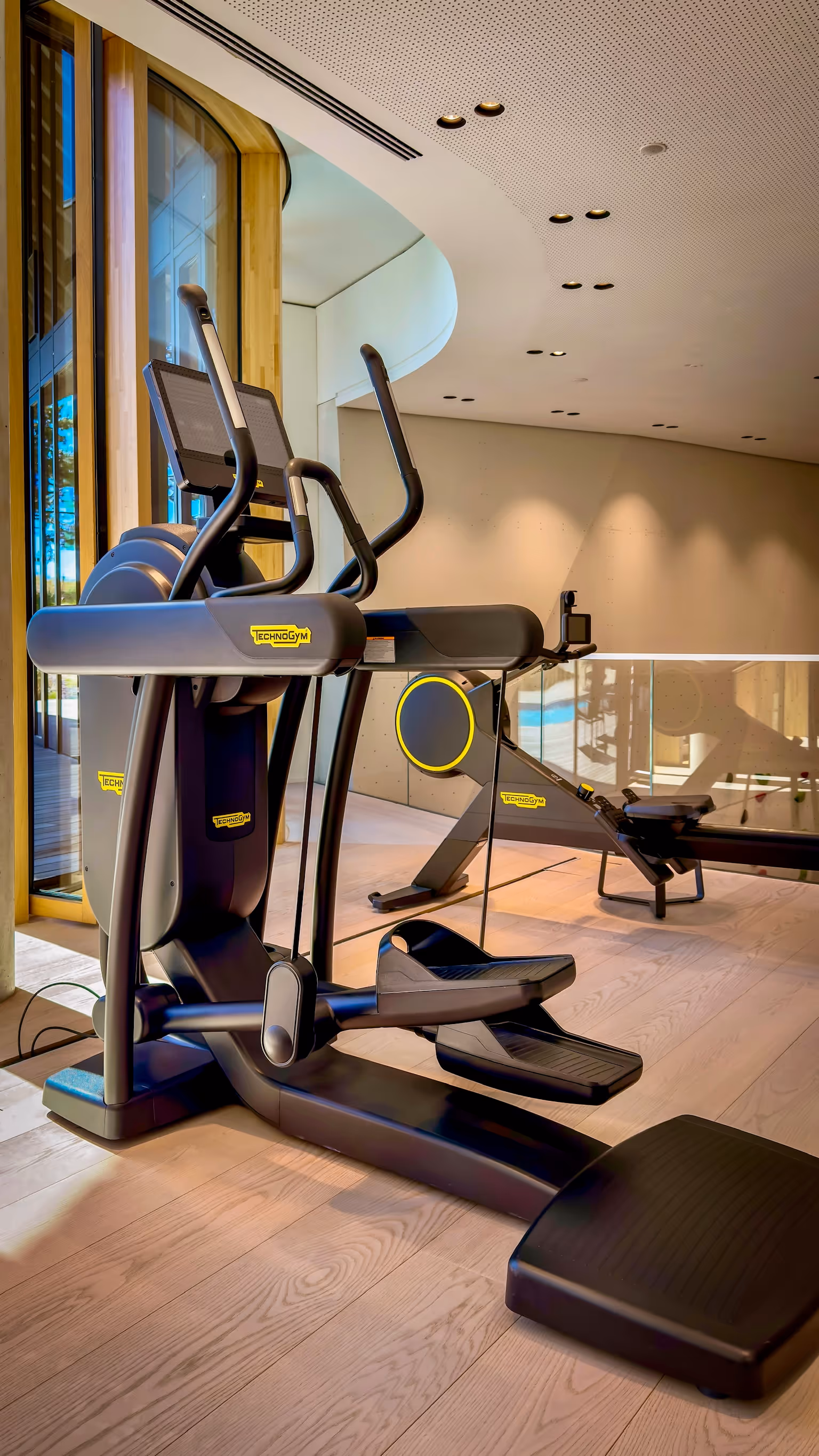 Modern elliptical machine and rowing machine in a bright fitness room with wooden floor and large windows.