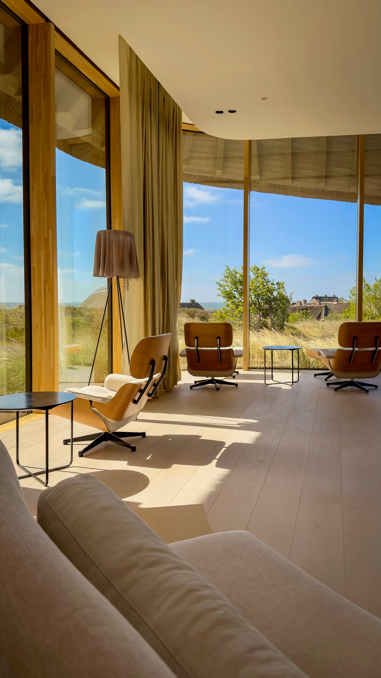 Bright room with large windows, modern chairs, side tables, lamp, and a view of greenery and houses.