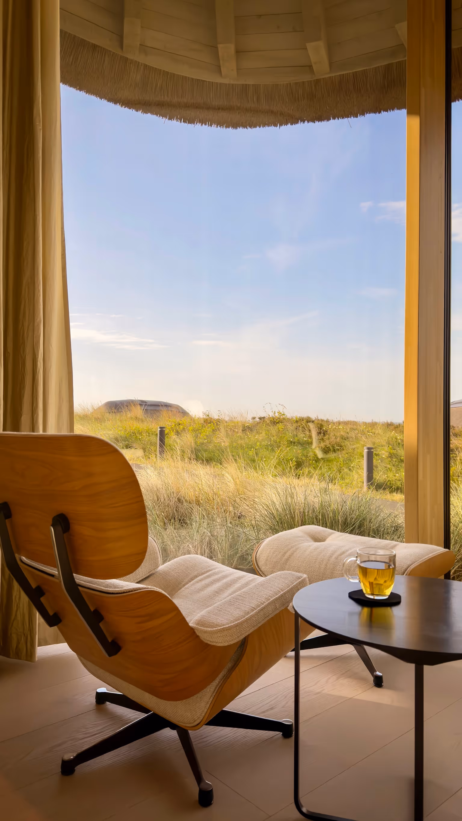 Modern lounge chair with ottoman by window overlooking grassy landscape and blue sky.