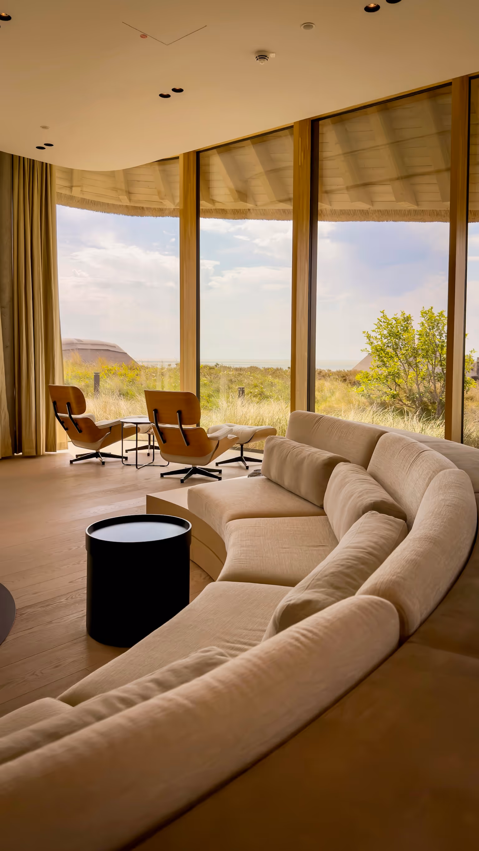 Modern living room with curved beige sofa, black round table, wood chairs, and large glass windows overlooking greenery.