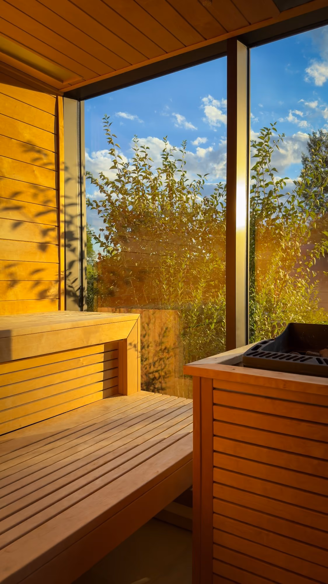 Wood-paneled sauna interior with bench and heater, overlooking green foliage and blue sky outside.
