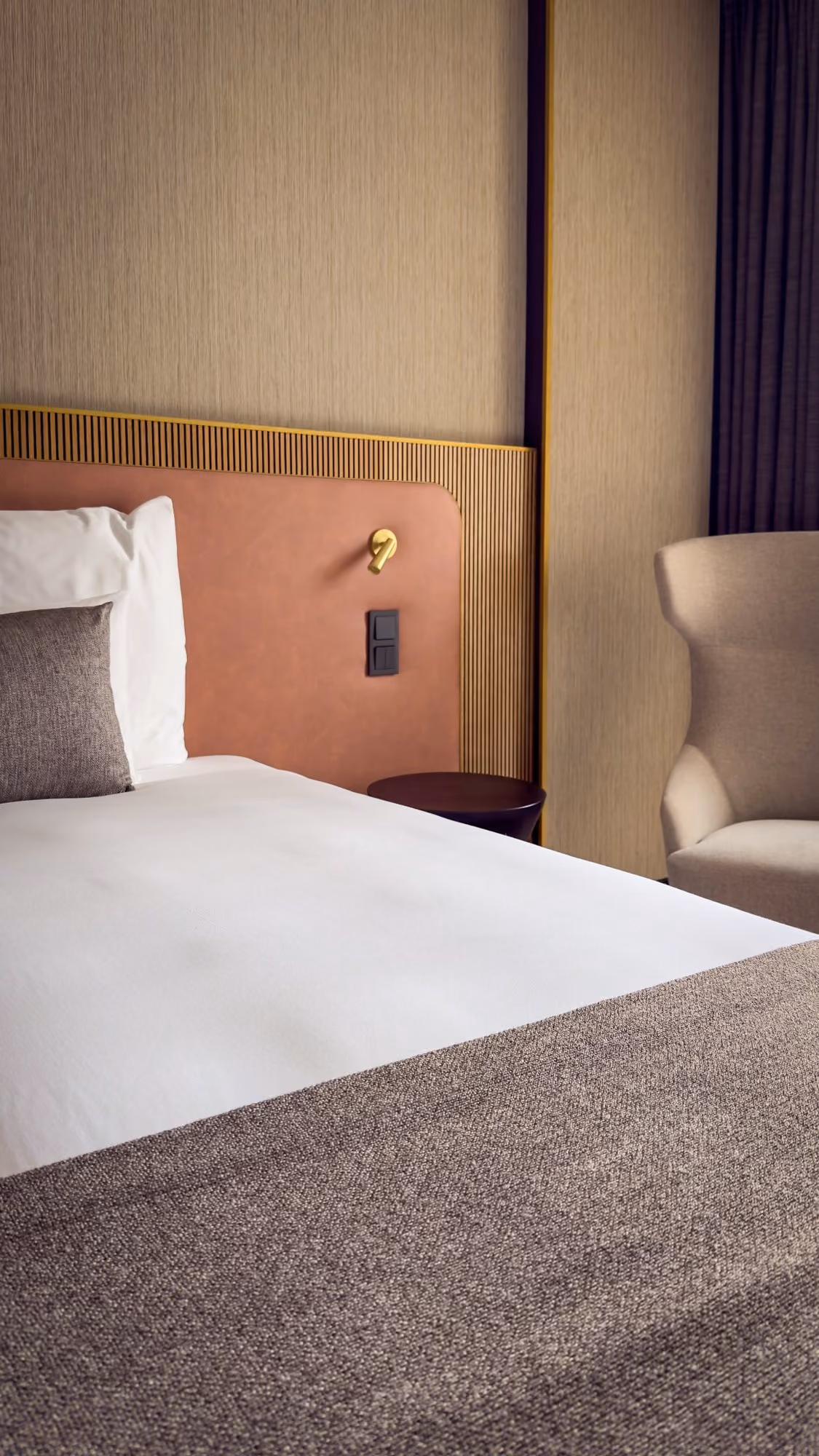 Modern hotel room corner with a white bed, gray pillow, beige chair, and textured brown headboard.