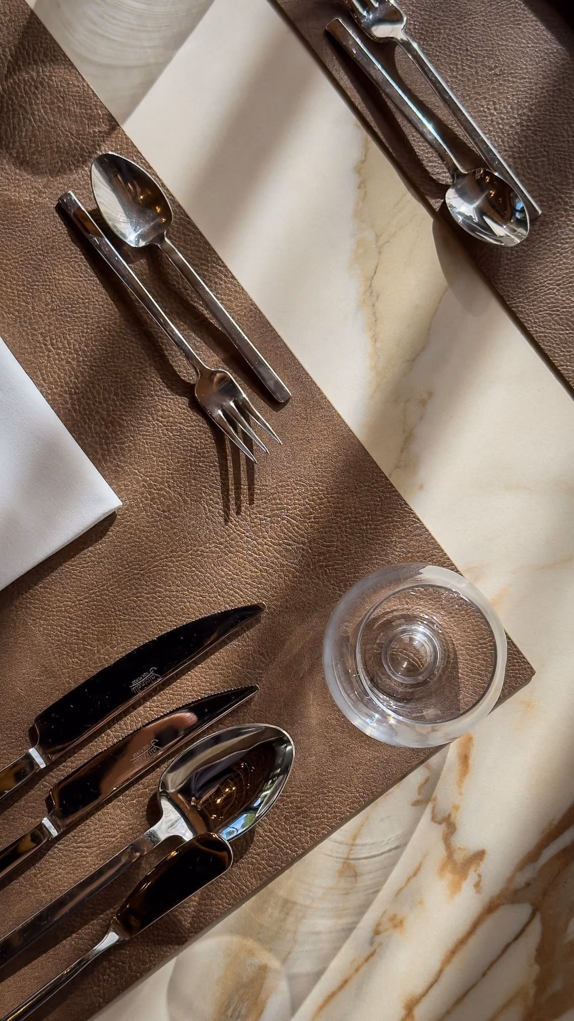 Table set with shiny stainless steel cutlery, a drinking glass, white napkin, and brown placemat.