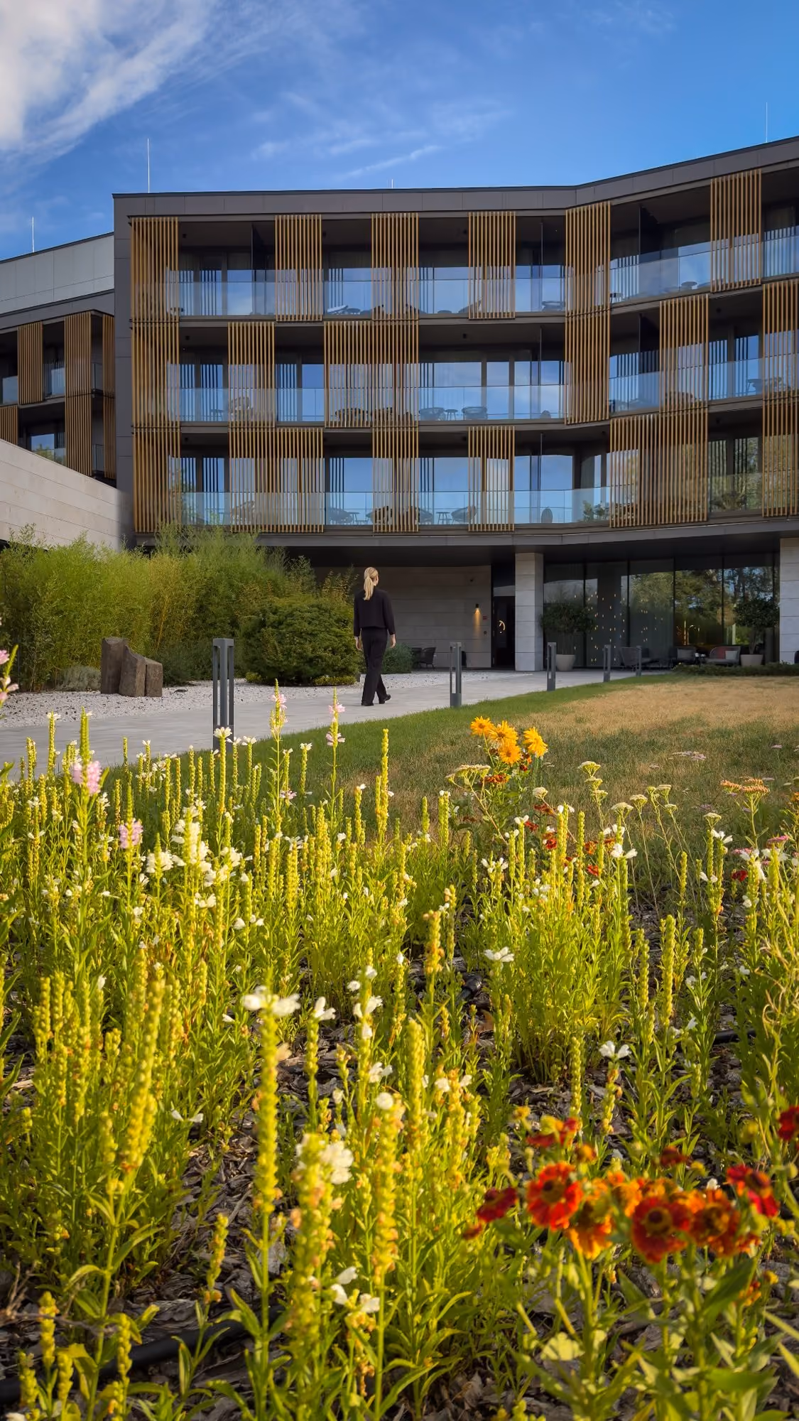 Woman walking on path in front of modern hotel with garden flowers and clear blue sky.