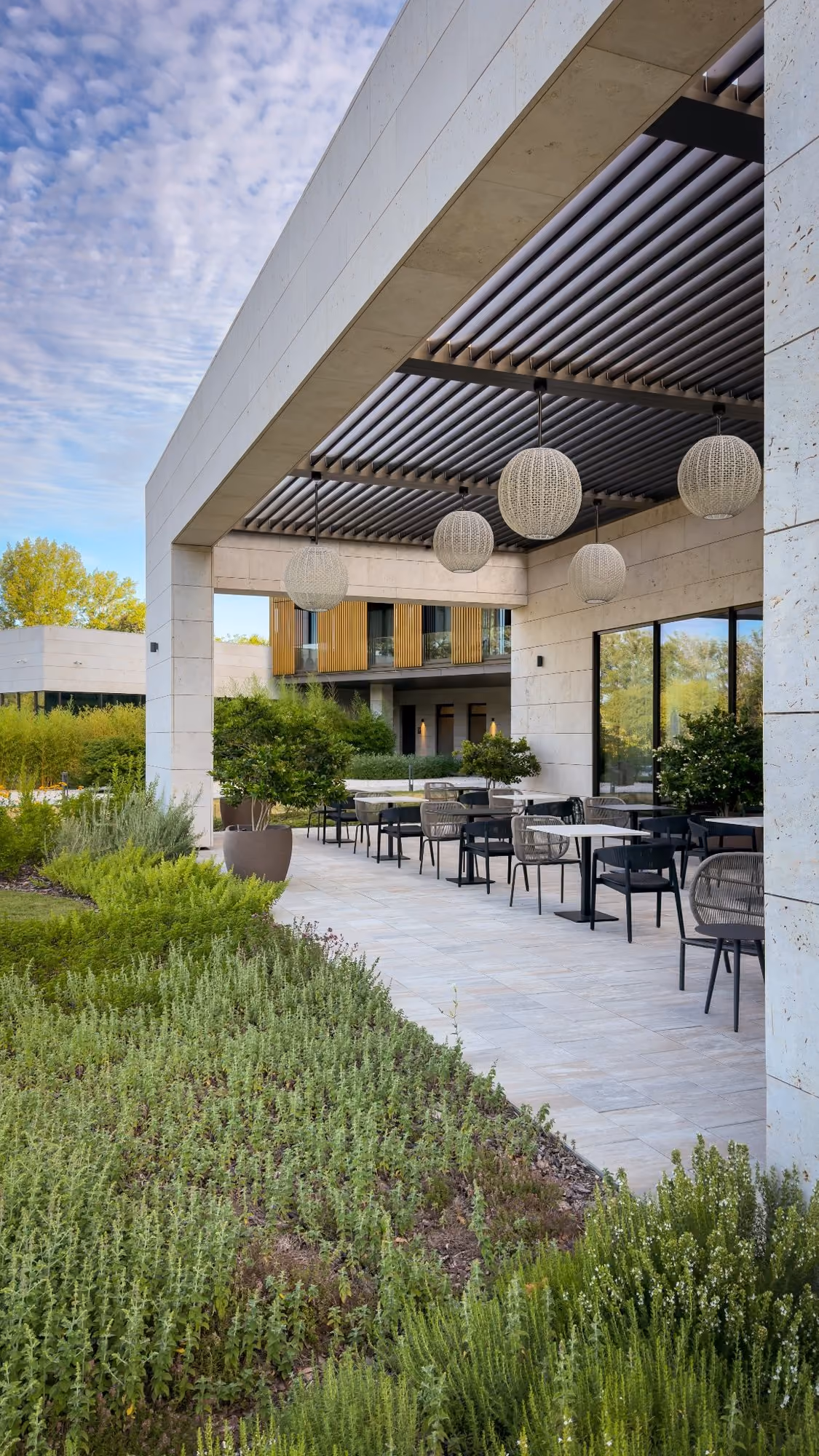 Modern hotel terrace with tables, chairs, hanging lights, and lush green landscaping under a partly cloudy sky.