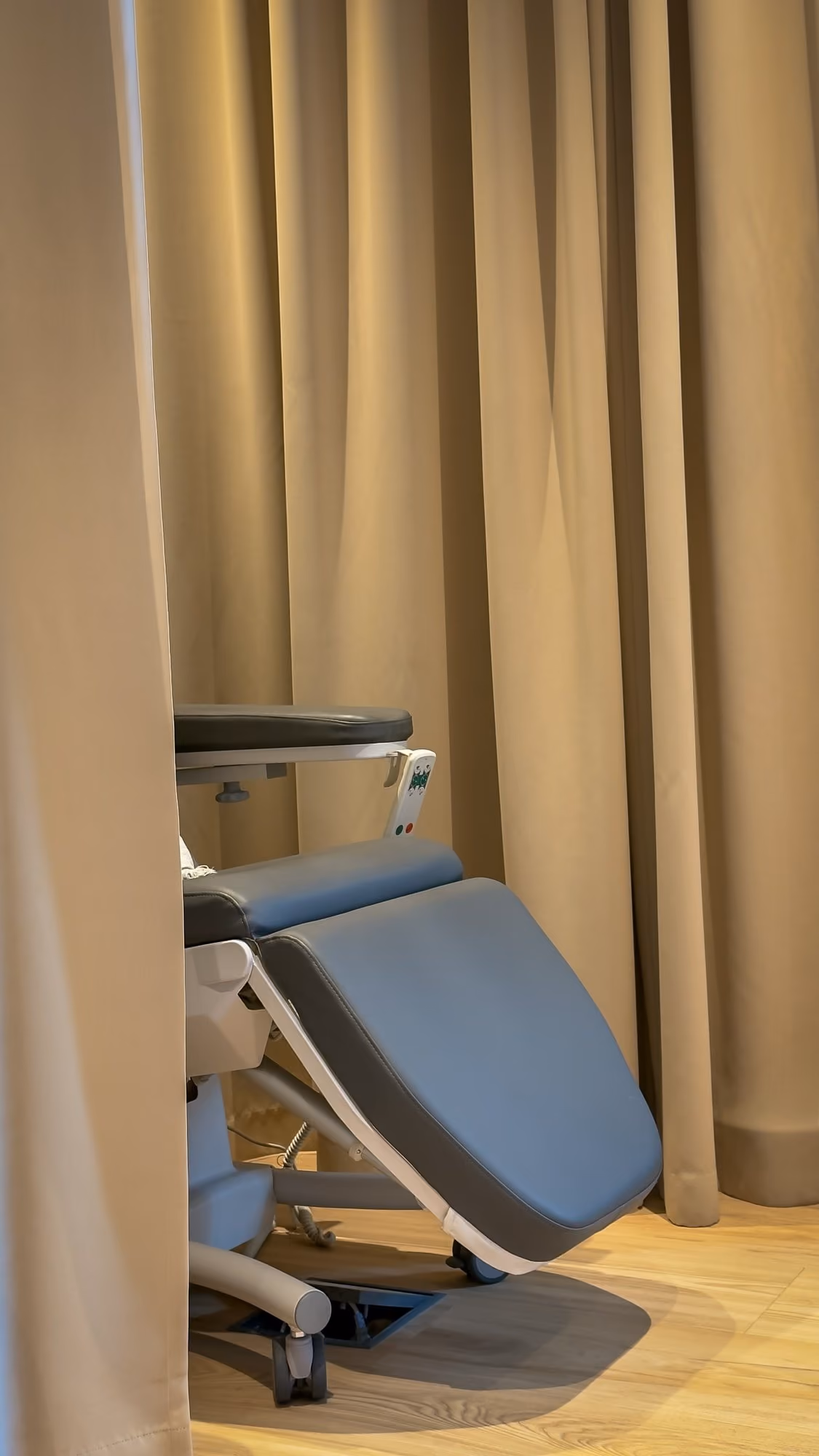 Side view of a medical or treatment chair with blue cushions behind beige curtains on wooden floor.