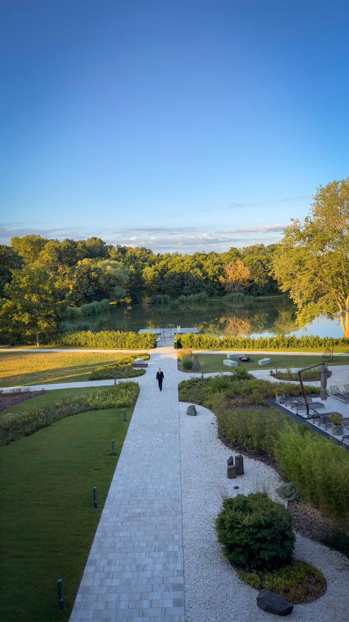 Person walking on paved path through green garden toward lake surrounded by trees under blue sky.