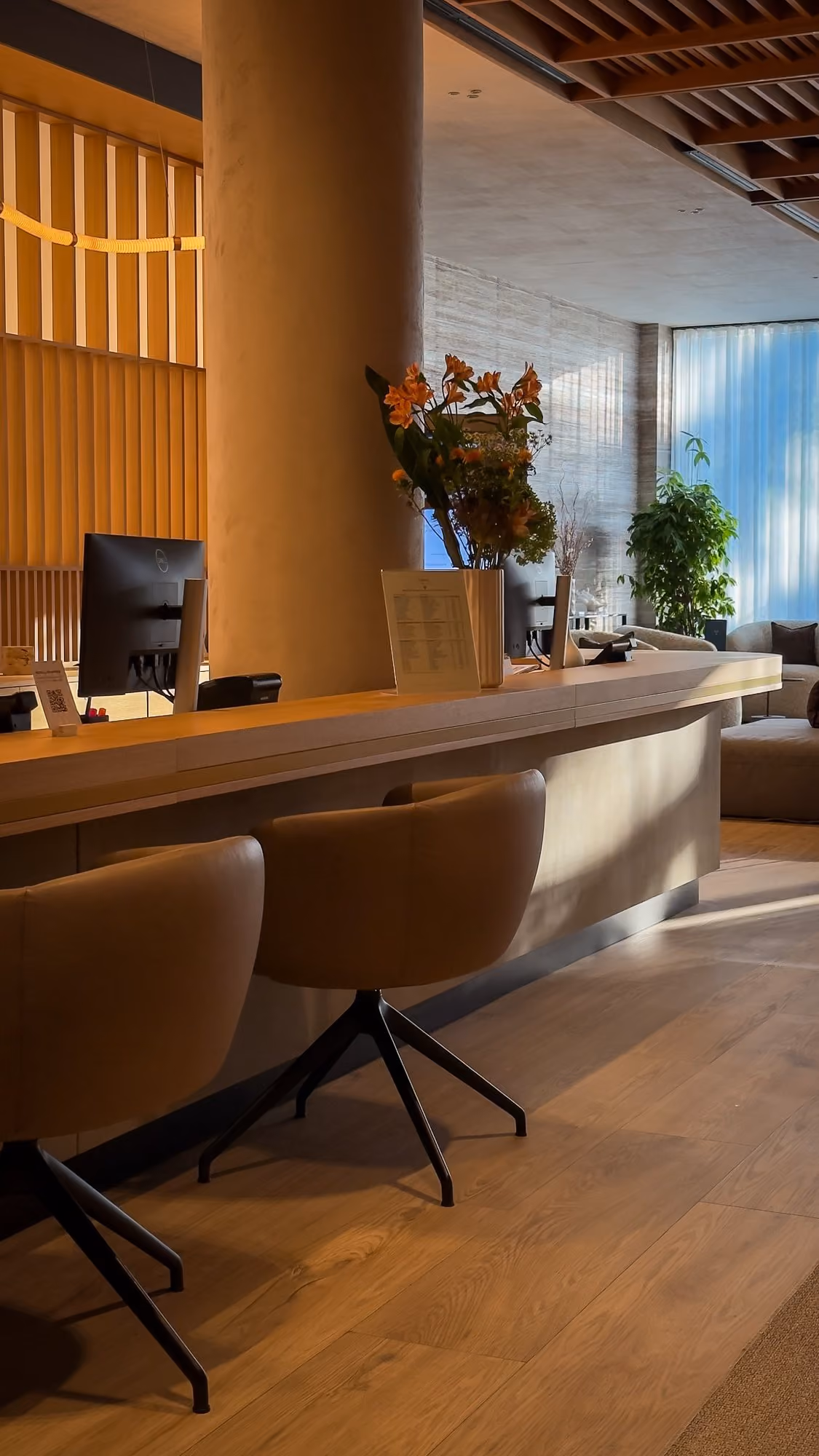 Modern reception area with wood desk, computer monitors, brown chairs, flowers, and plants by window.