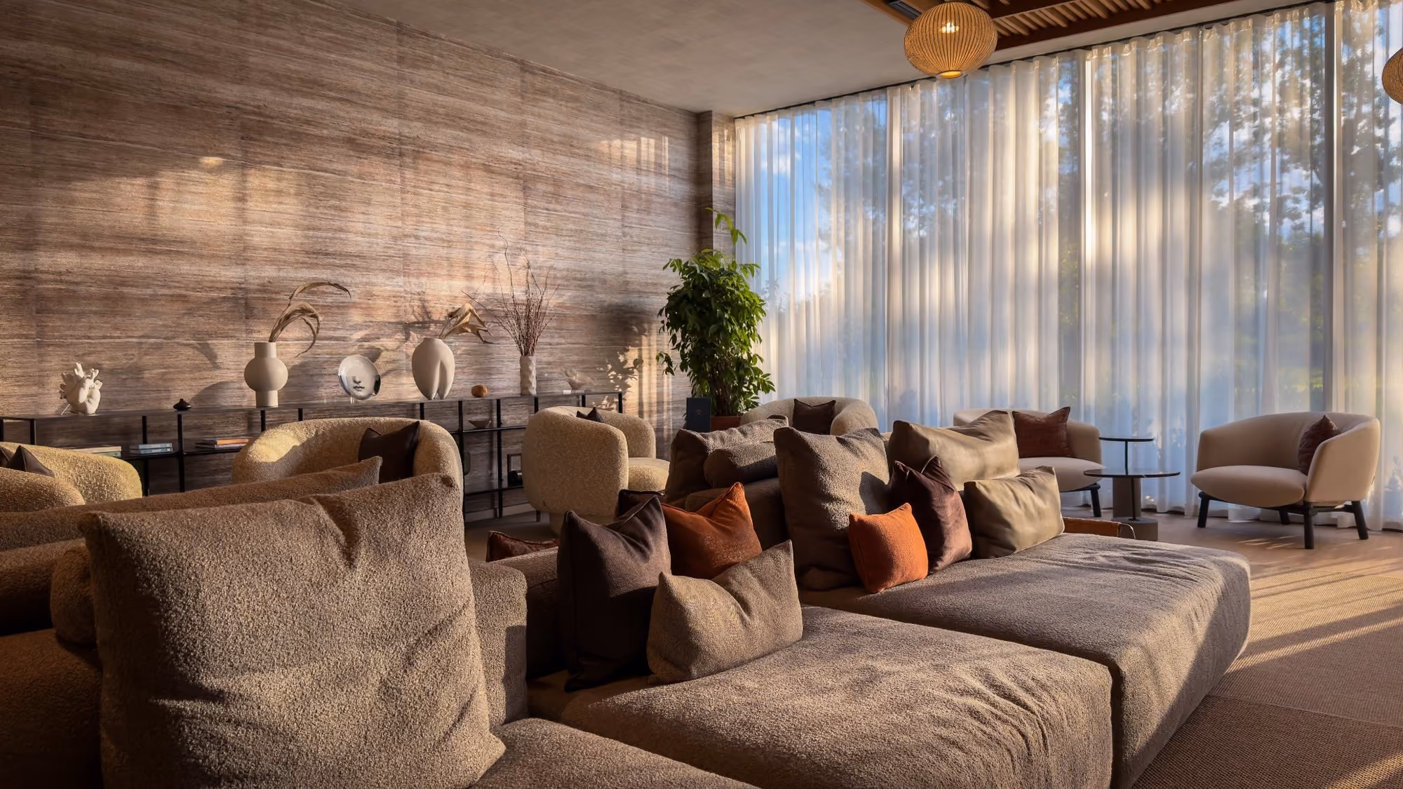 Cozy lounge with beige sofas, armchairs, cushions, potted plant, decorative shelf, and sheer curtains.