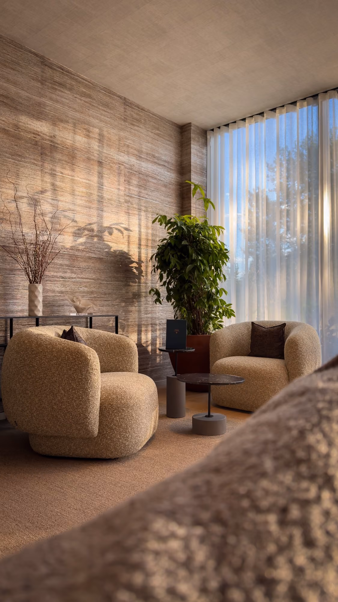 Modern lounge with two beige armchairs, a potted plant, side tables, and sunlight through sheer curtains.