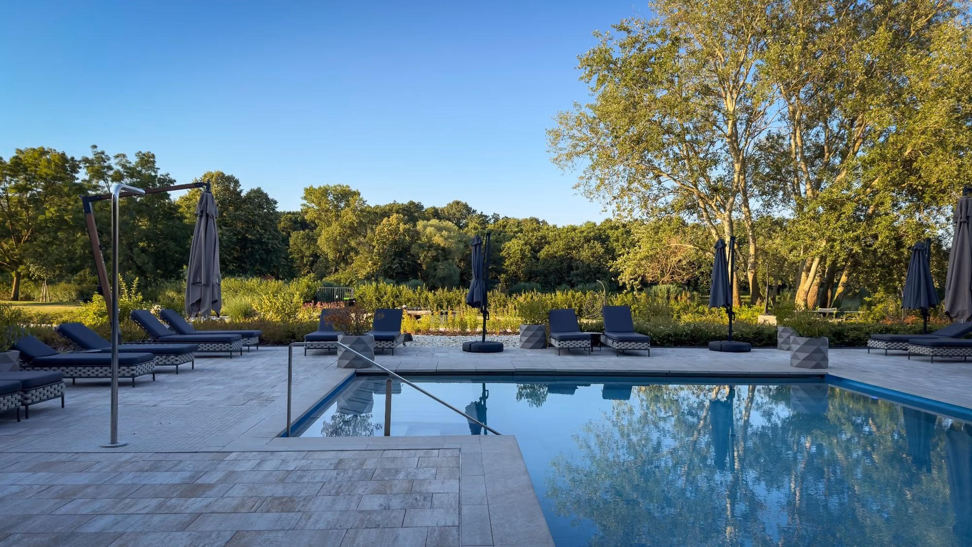 Outdoor swimming pool with blue lounge chairs and umbrellas set around it, trees in the background.