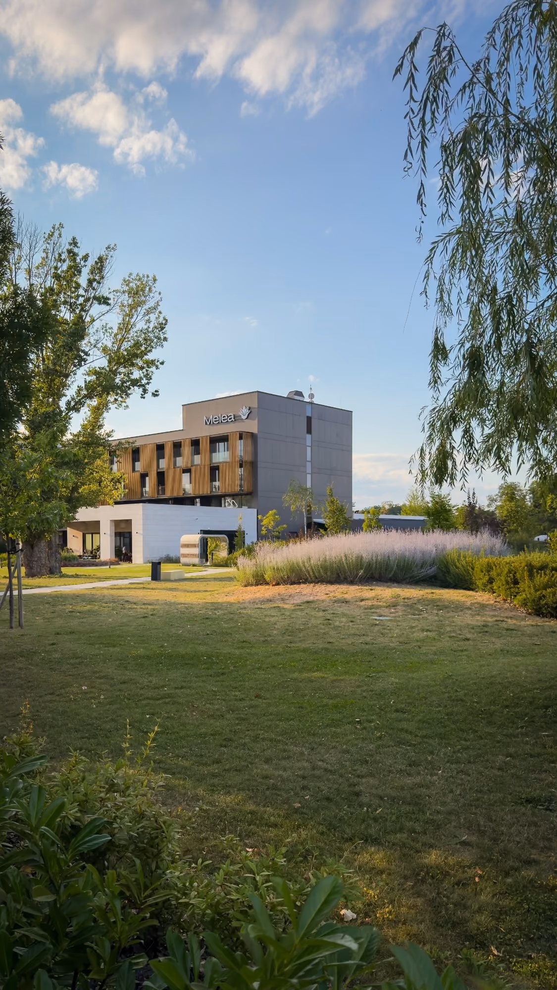 Modern building named Melea surrounded by green lawn, trees, and bushes under a blue sky with clouds.