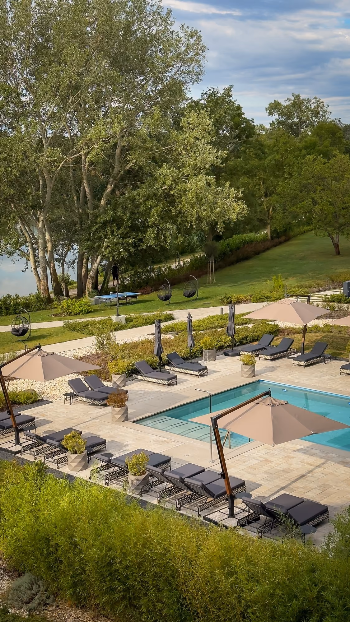 Outdoor pool with a small arched wooden bridge, surrounded by lounge chairs, greenery, and a hotel building with mountains in the background under clear blue sky.